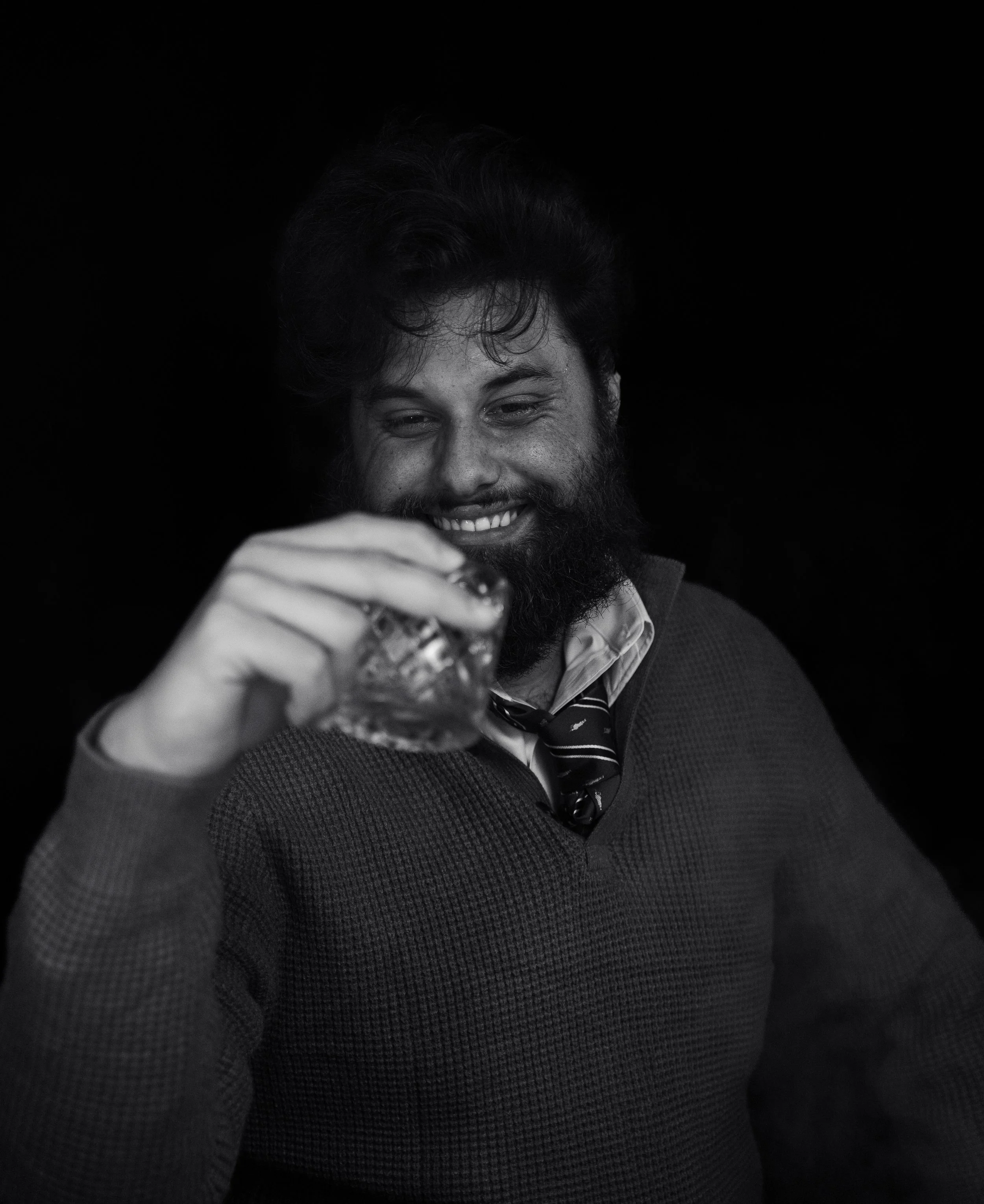 A man with dark hair and a beard smiling and holding a glass of drink, wearing a suit with a tie, against a dark background.