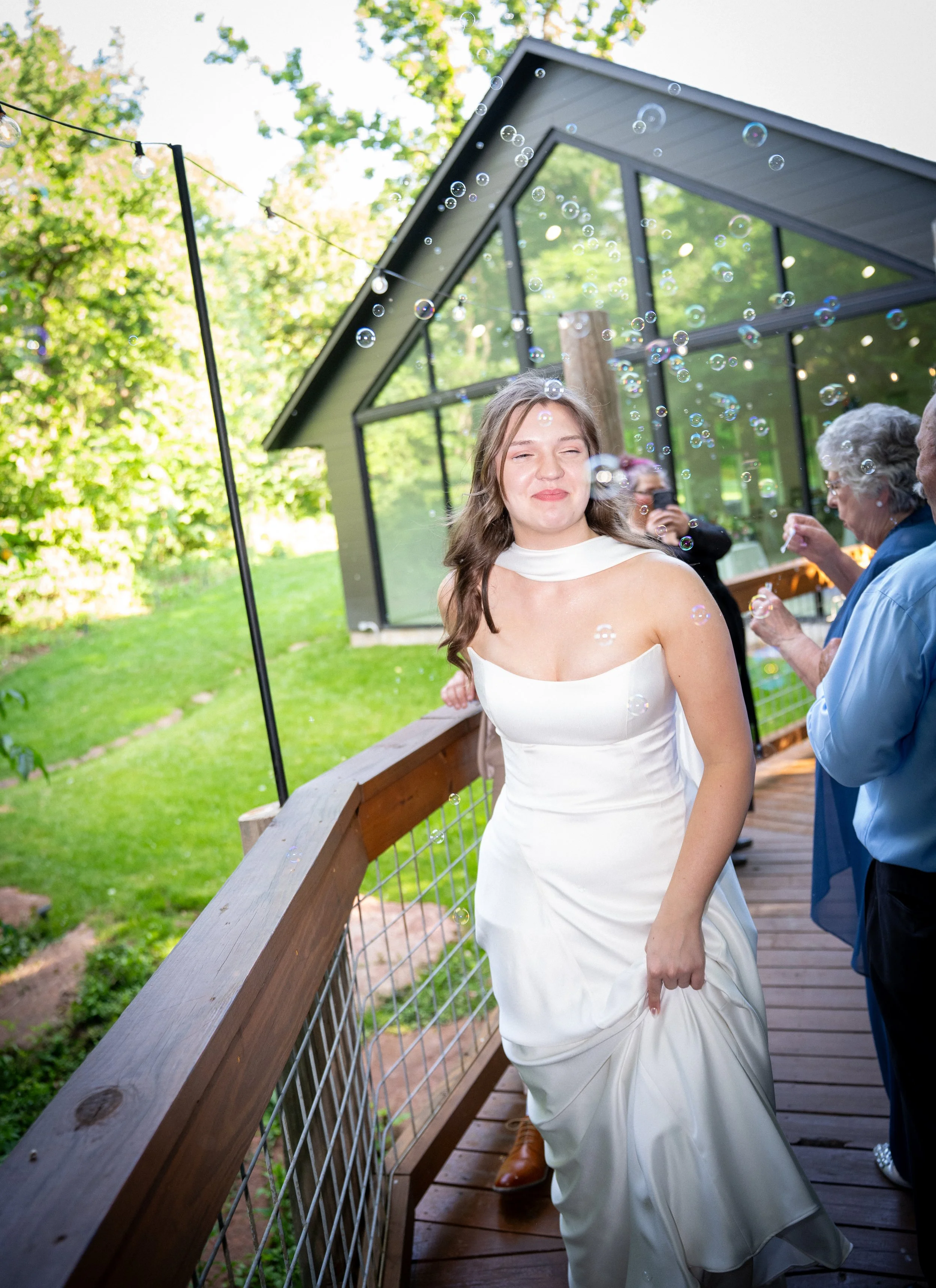 A young woman in a white satin wedding dress is standing on a wooden deck outdoors, smiling with her eyes closed. Bubbles are floating in the air around her. In the background, there are older women and a modern glass building surrounded by trees and