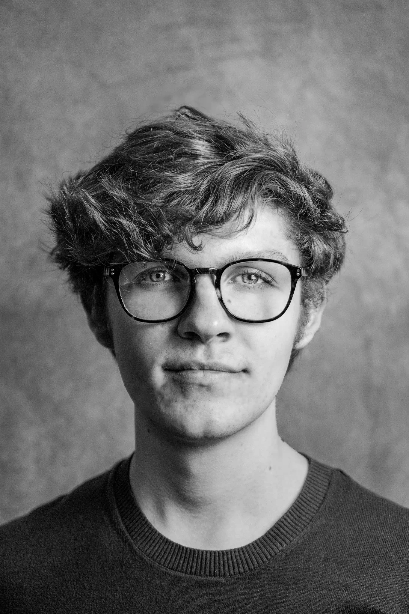 Black and white photo of a young person with short, wavy hair and glasses, wearing a dark sweater, smiling slightly, against a plain background.