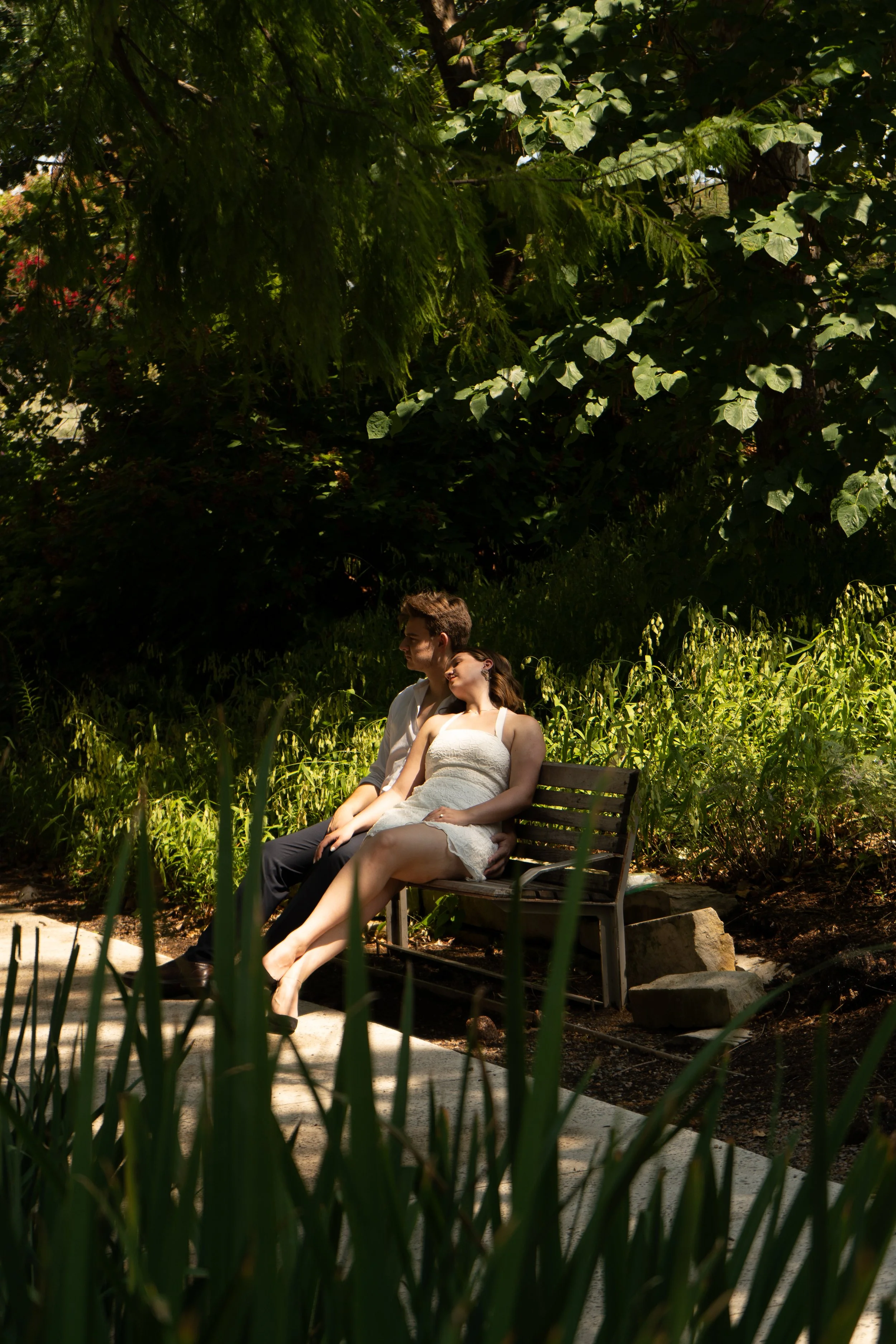 A couple sitting closely together on a park bench in a lush green garden with dense foliage overhead and plants at the foreground.