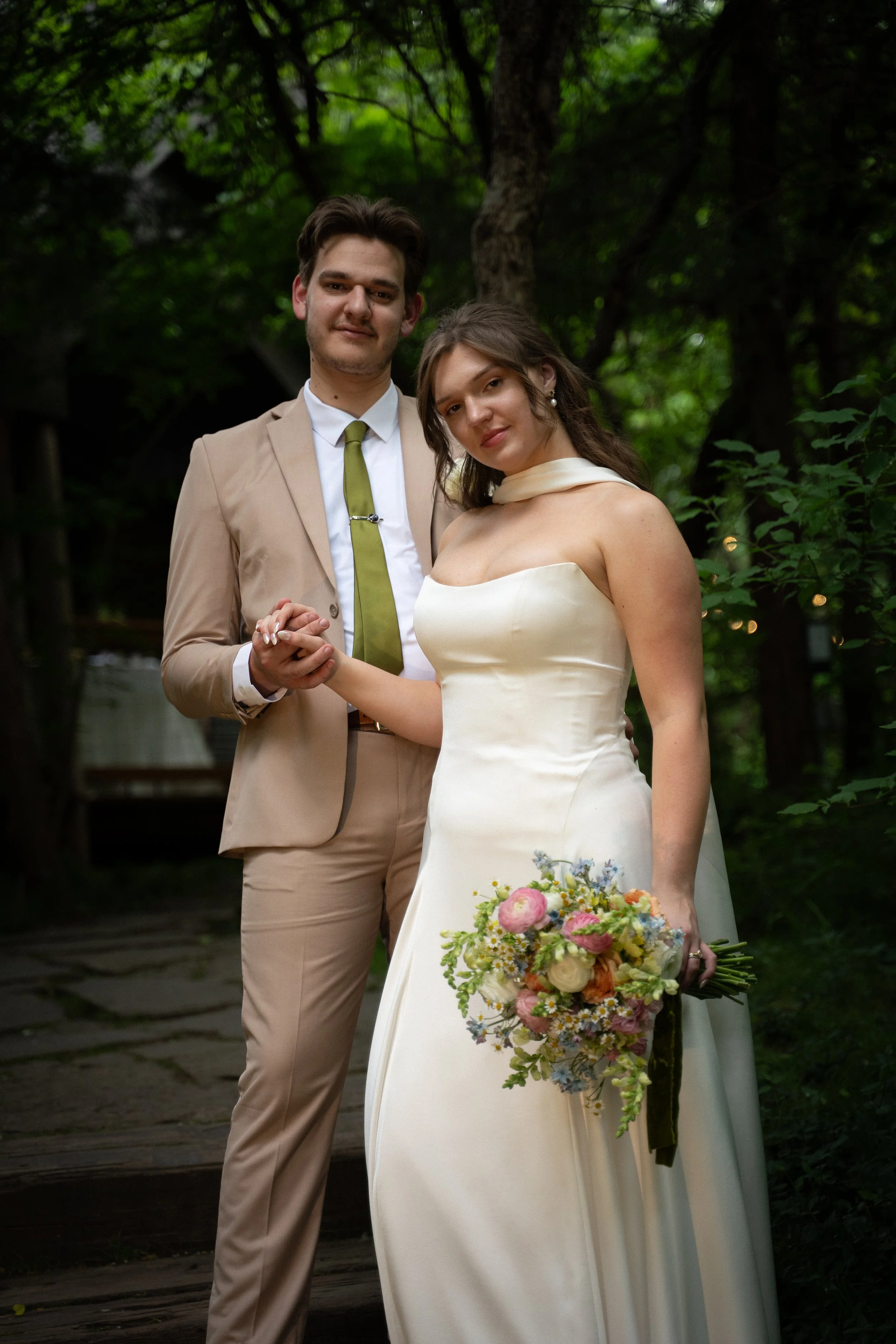 A young couple dressed in wedding attire holding hands outdoors surrounded by green trees. The woman is wearing a white wedding dress and holding a colorful bouquet, while the man is dressed in a beige suit with a white shirt and a green tie.