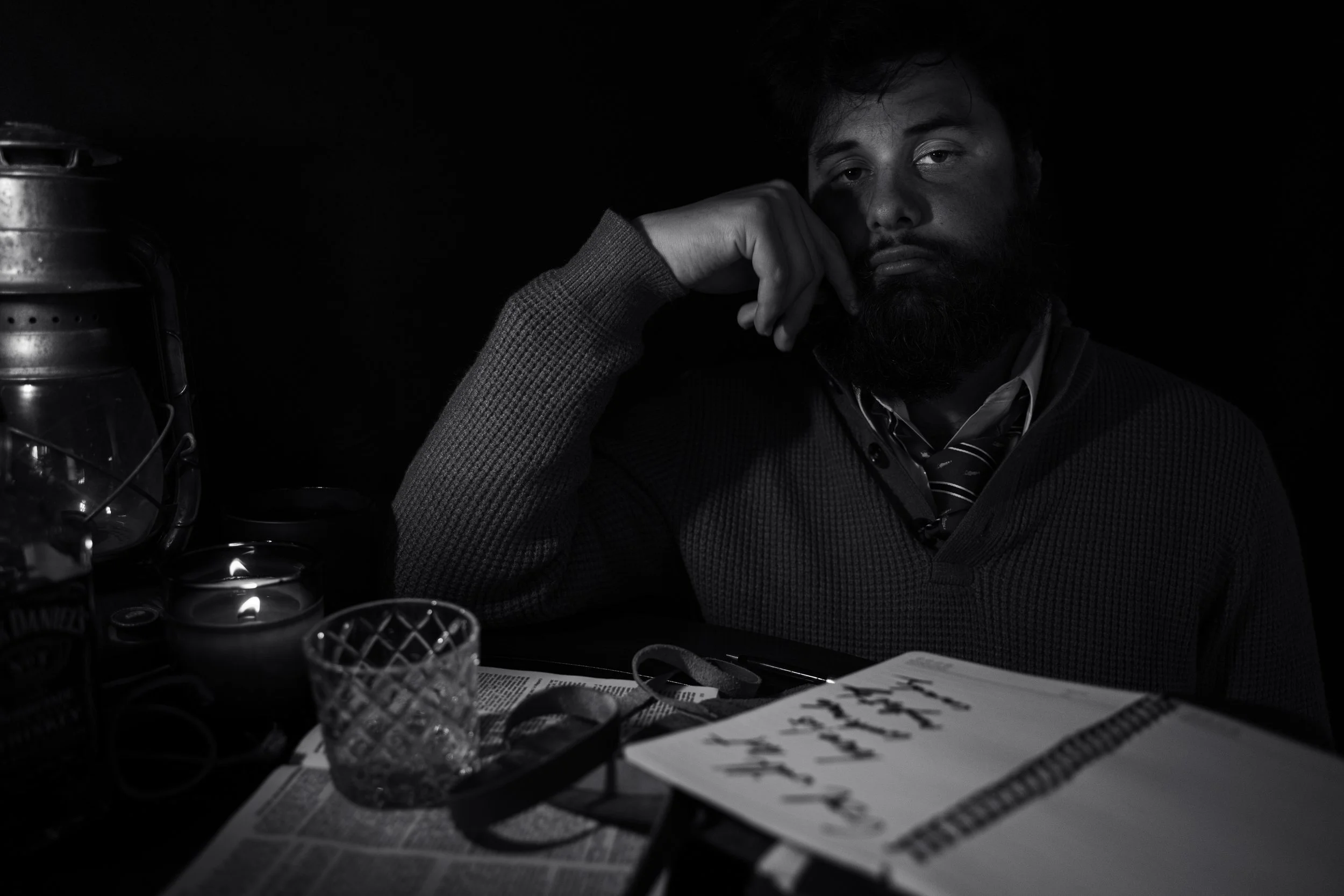 A man with dark hair and a beard resting his head on his hand, sitting at a table with newspapers, glasses, and a lantern, in a dimly lit environment.