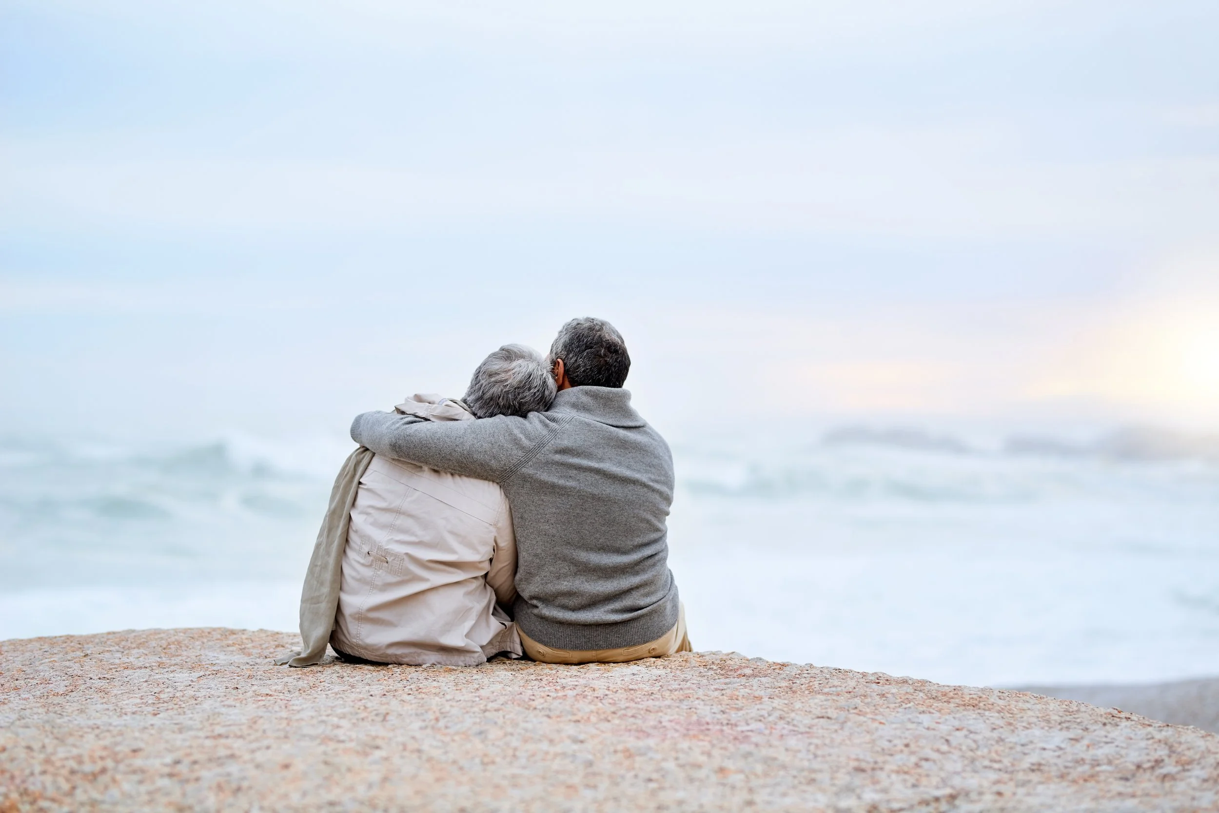An elderly couple sitting on a rocky beach, embracing and looking at the ocean.