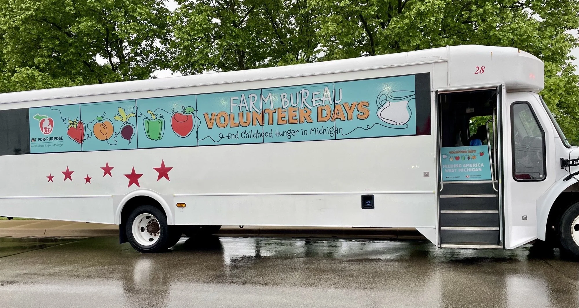 A white bus with a colorful banner on its side that reads "Farm Bureau Volunteer Days" and "End Childhood Hunger in Michigan." The banner features illustrations of various vegetables and a coffee mug. There are red stars on the lower part of the bus, and the bus is parked on a wet surface with green trees in the background.