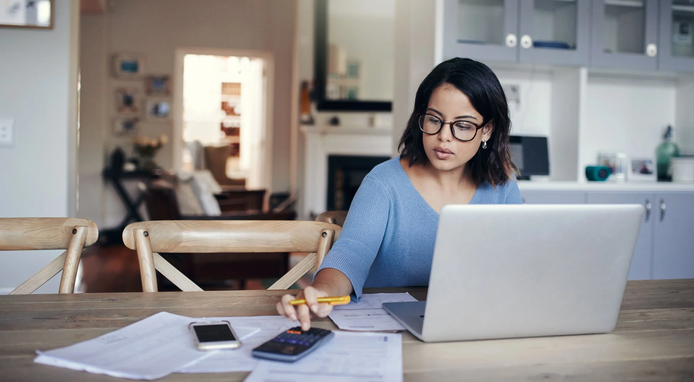 A young woman with dark hair and glasses working at a cluttered wooden table with papers, a smartphone, and a calculator, using a laptop in a kitchen.