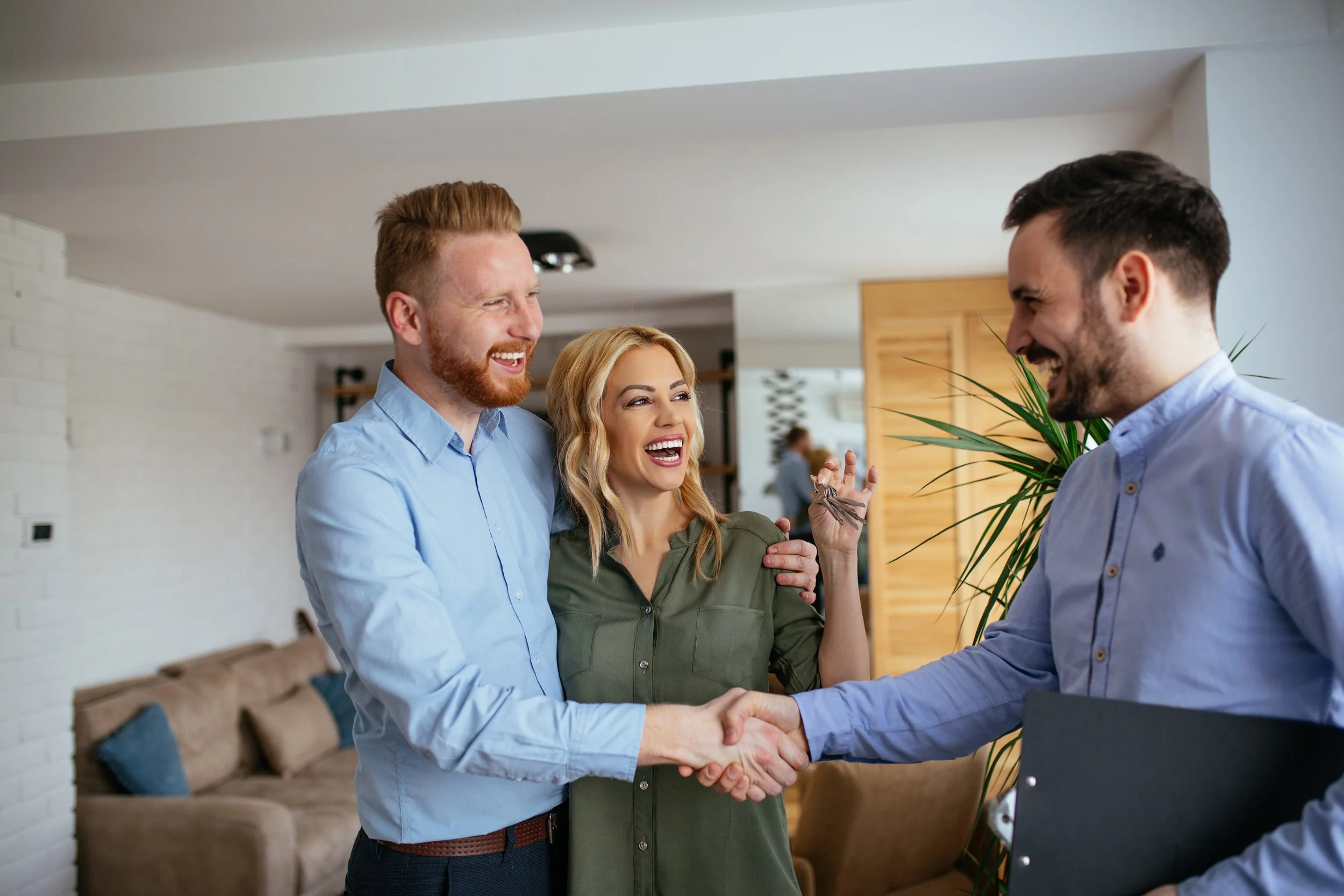 Three people, two men and a woman, smiling and shaking hands in a living room. The woman is wearing an olive green top, the man on the left has red hair and a beard, and the man on the right has dark hair and a beard, dressed in a light blue shirt.