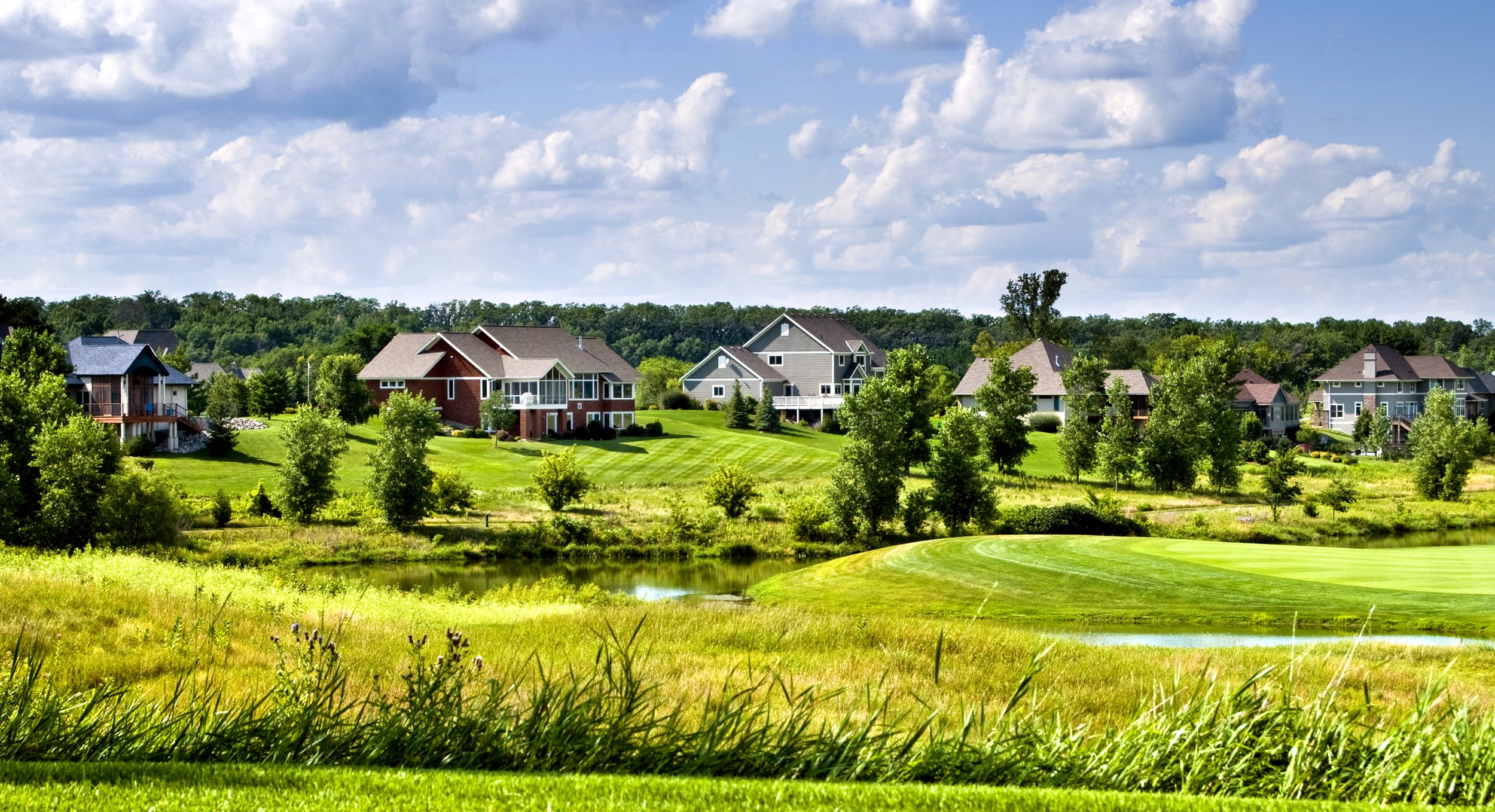 Suburban neighborhood with large houses, green lawns, trees, a small pond, and a blue sky with clouds.