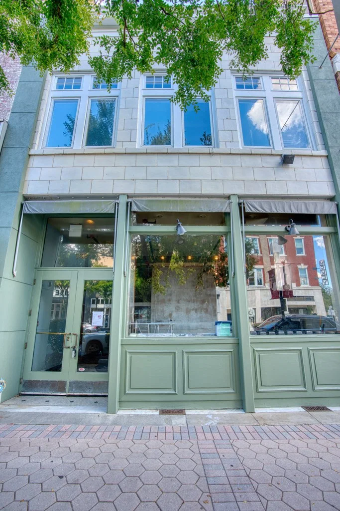 Empty storefront with green trim and large window display, upper floors with three sets of windows, and a tree partially covering the top of the building.