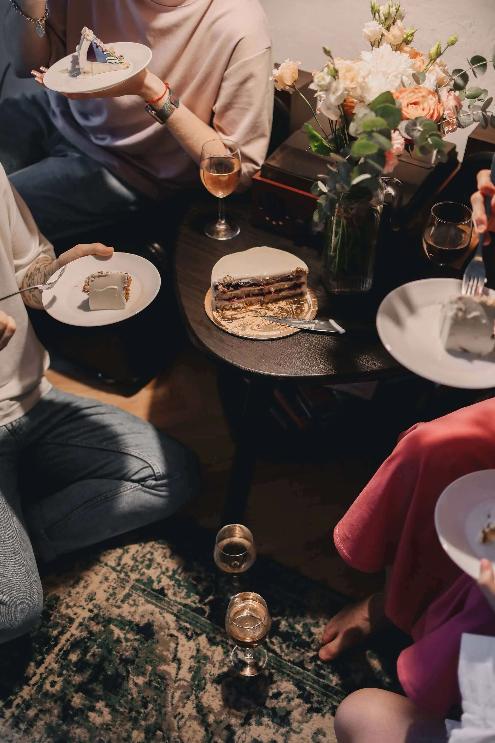 People gathered around a small table with cake slices, glasses of rosé wine, and a vase of flowers, celebrating together in a cozy indoor setting.