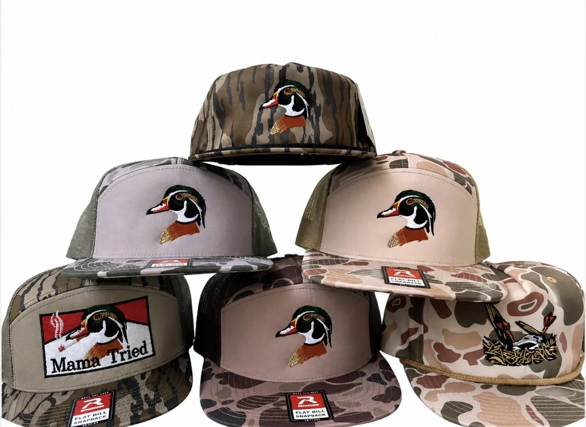 Stacked camouflage-patterned baseball caps featuring a duck head embroidery and a patch that says "Mama Tried" on a camouflage hat.