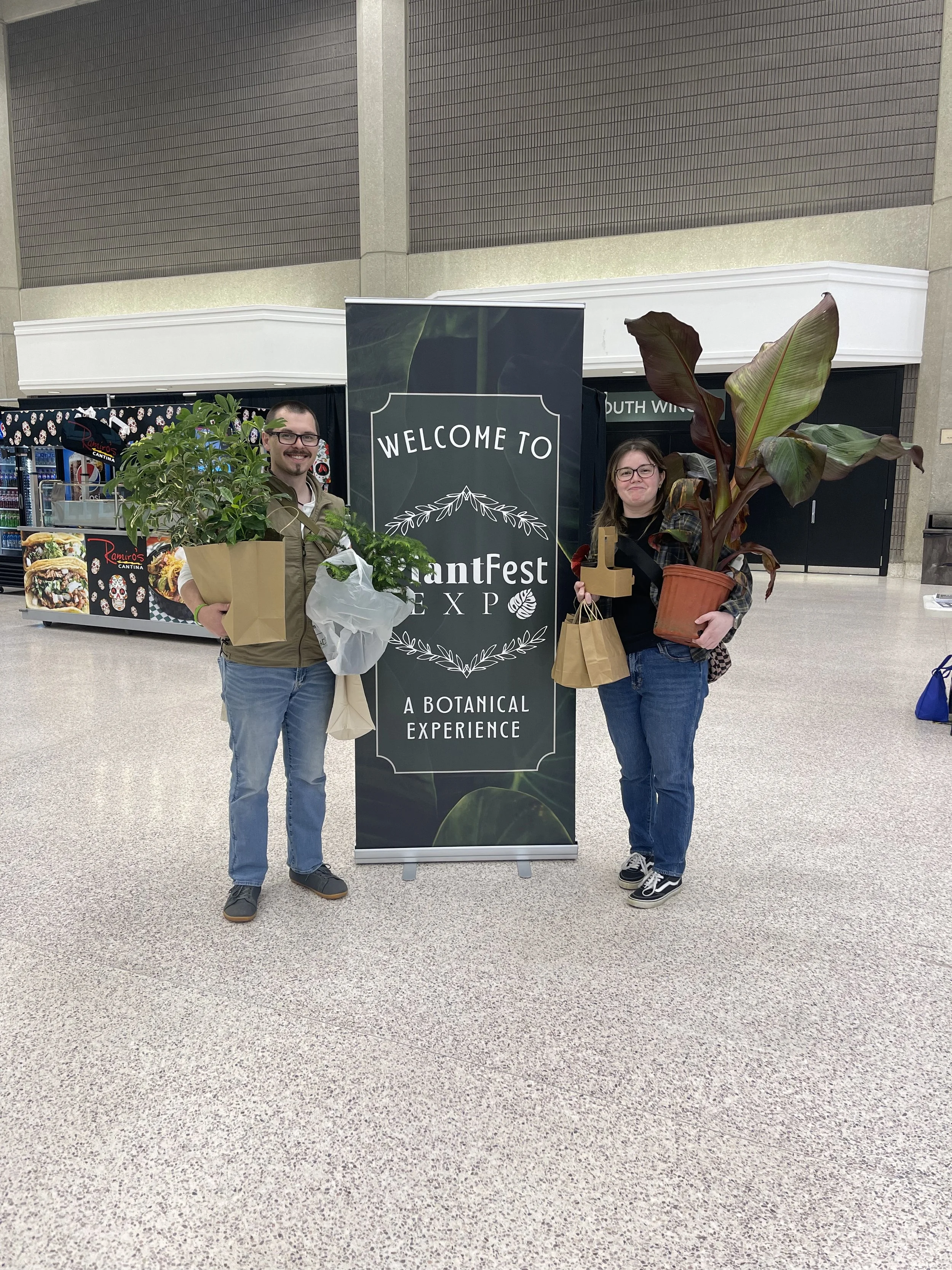 Two people standing in front of a sign at an event, holding plants and shopping bags.