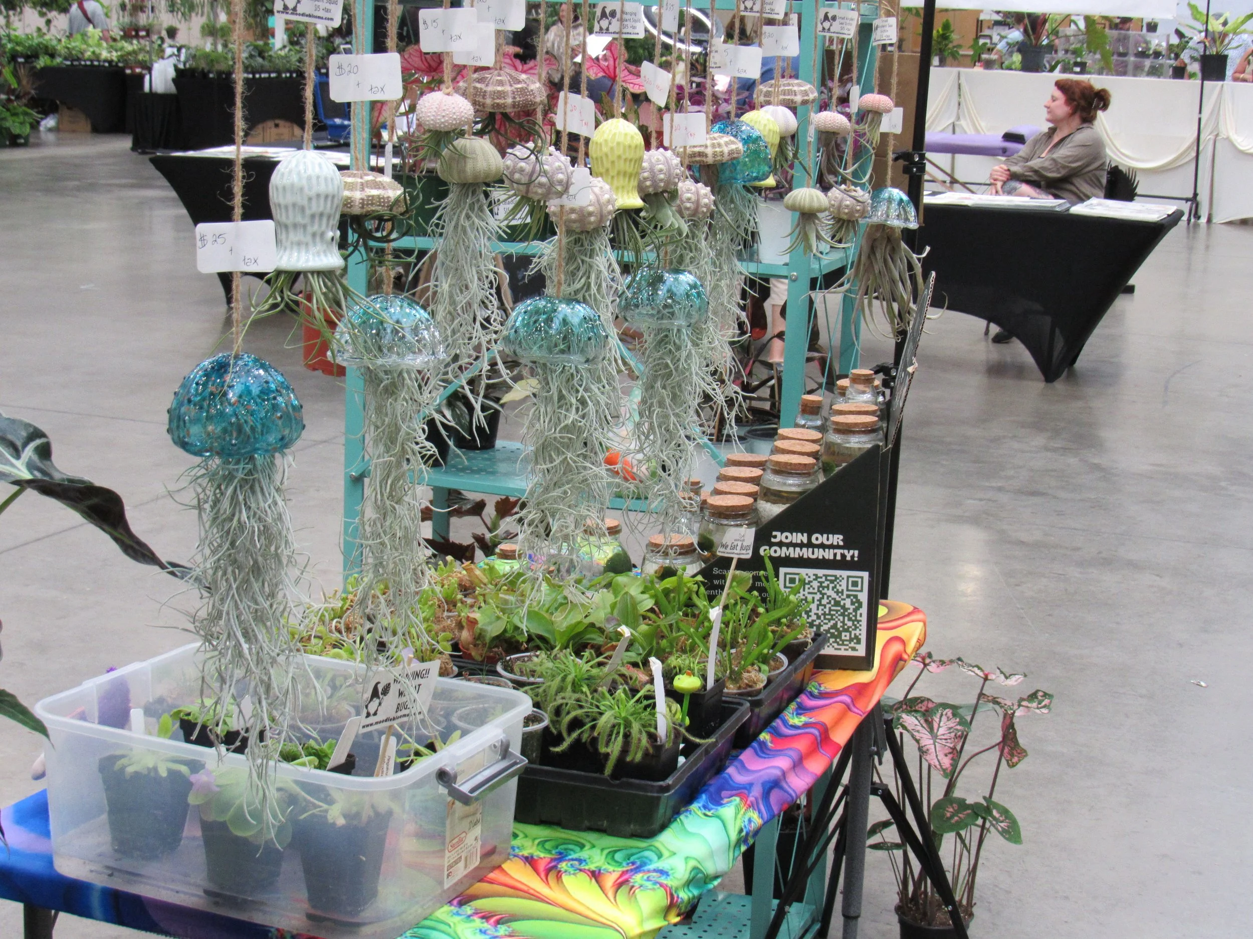 A display of handmade ceramic jellyfish and other decorative items at an indoor market. The display includes hanging jellyfish with detailed tentacles and glowing blue bell-shaped bodies, various potted plants, and signs with prices. A woman with red