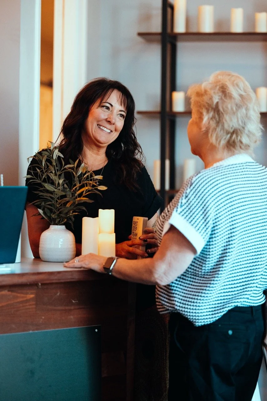 Two women engaging in conversation at a reception desk, with candles and a plant on the counter.
