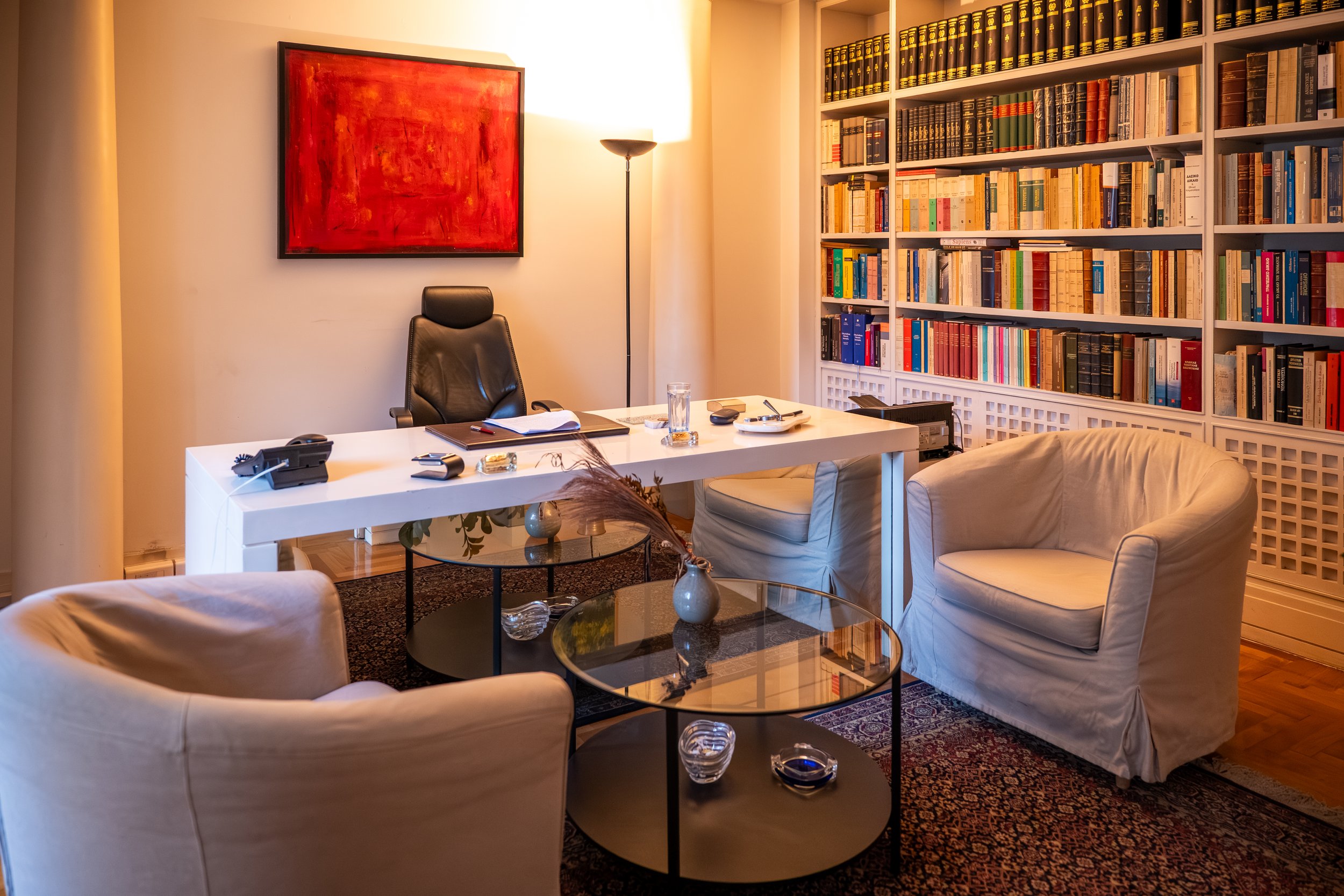 A cozy office with a white desk, black leather chair, and beige armchairs. There is a large bookshelf filled with books, a painting with red and orange hues on the wall, and a tall floor lamp providing warm lighting.