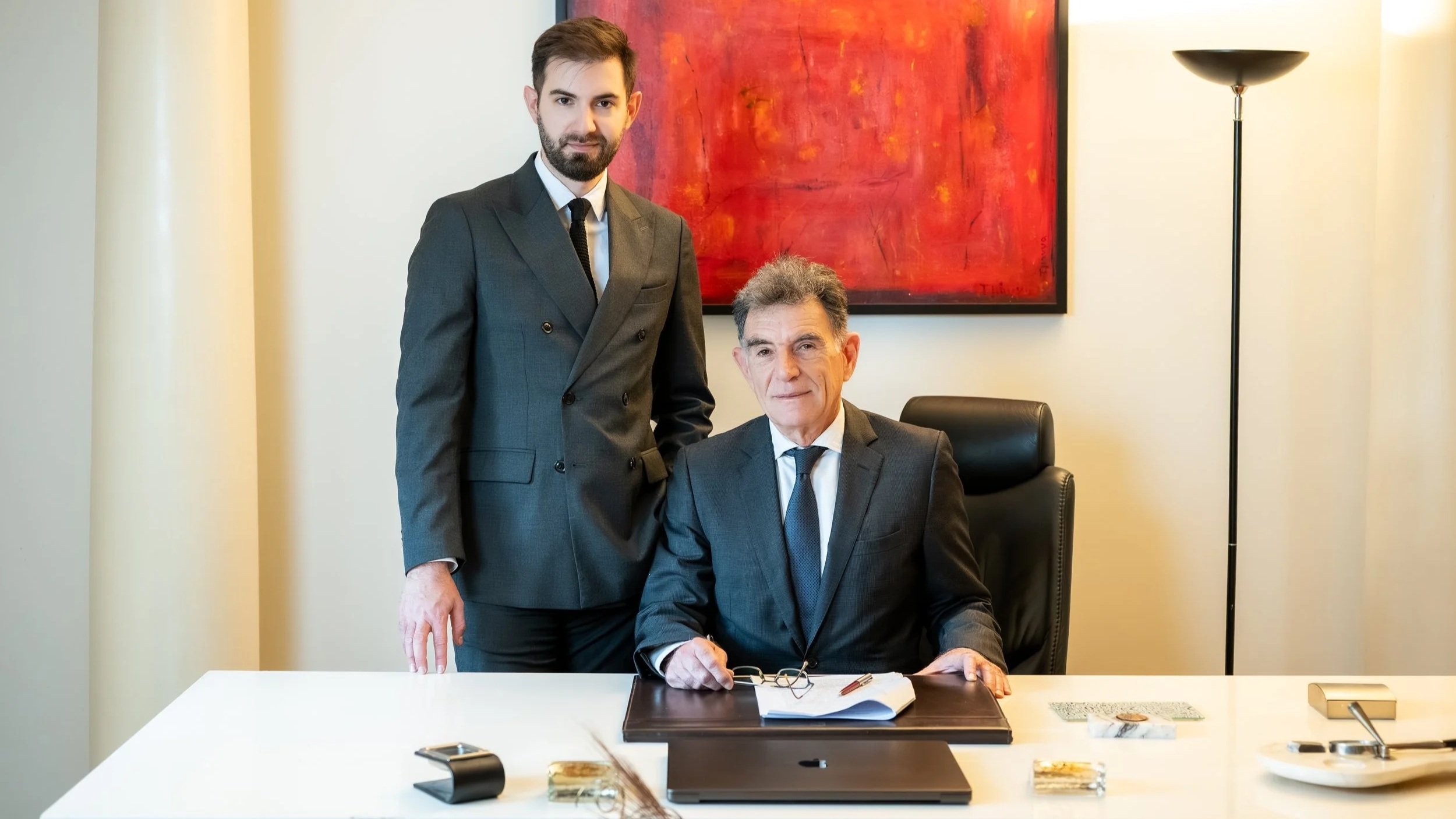Two men in business attire in an office. One is standing, and the other is sitting at a desk with office supplies and documents, with a large red abstract painting in the background.