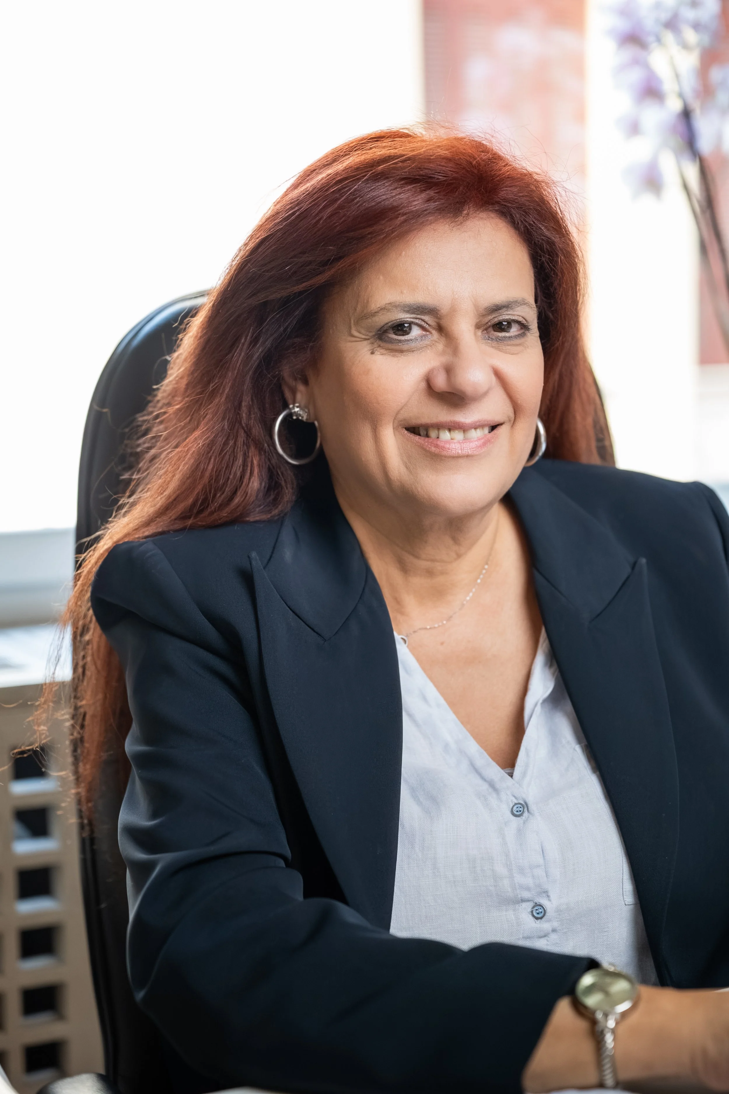 A middle-aged woman with reddish-brown hair, wearing a dark blazer, sitting at a desk in an office setting, smiling at the camera. She is accessorized with hoop earrings and a watch.