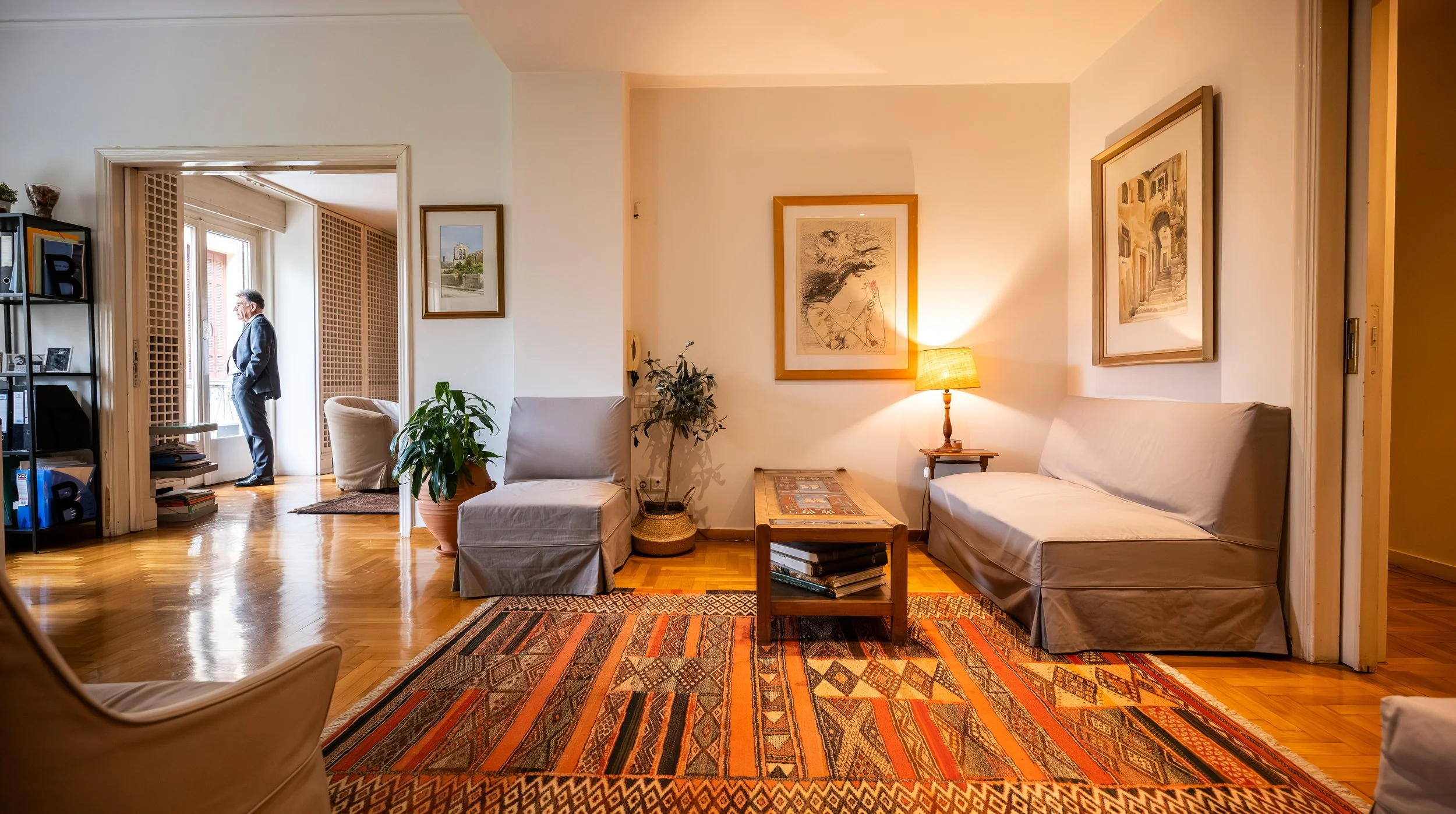 A cozy living room with a wooden floor, white walls, and framed artwork. There is a sitting area with a white sofa and a chair, a small coffee table with books, a lamp on a side table, potted plants, and a colorful rug. A man in a suit stands in a do