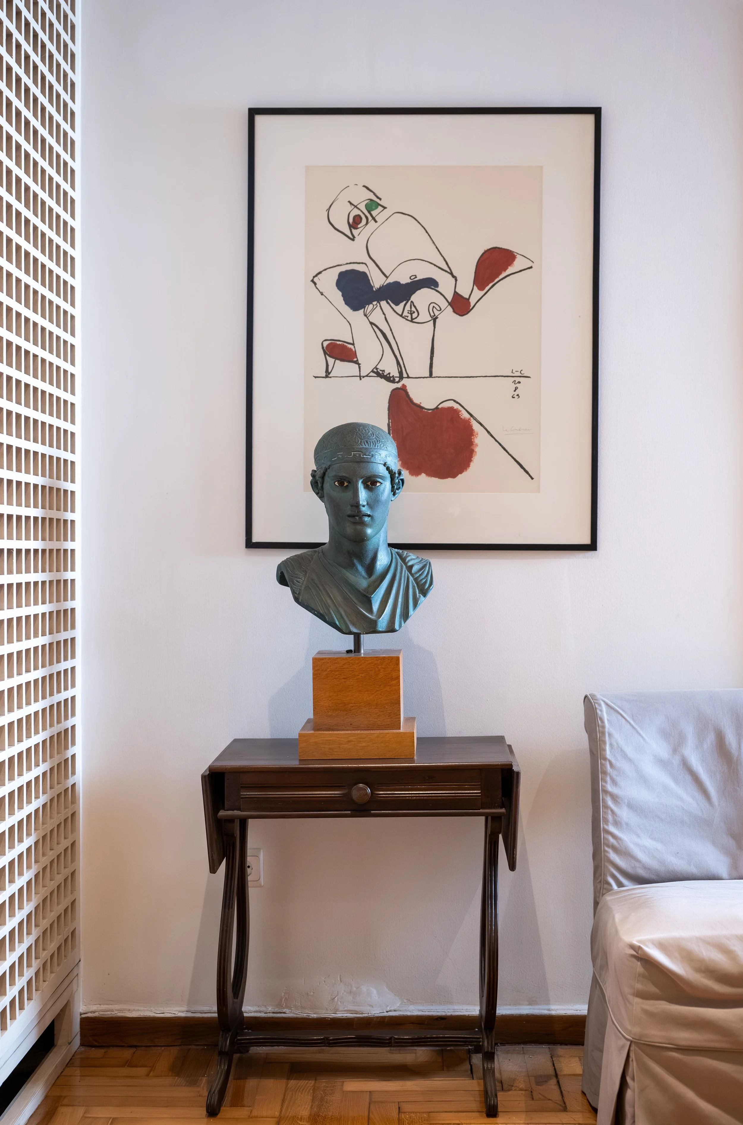 A decorative bust sculpture of a woman with classical style, painted in a greenish hue, placed on a wooden pedestal on top of a vintage wooden table. Behind it is a framed abstract artwork on the wall, featuring black lines and splashes of color, including red, black, and green. To the right, part of a beige armchair is visible, and on the left, a white lattice room divider.