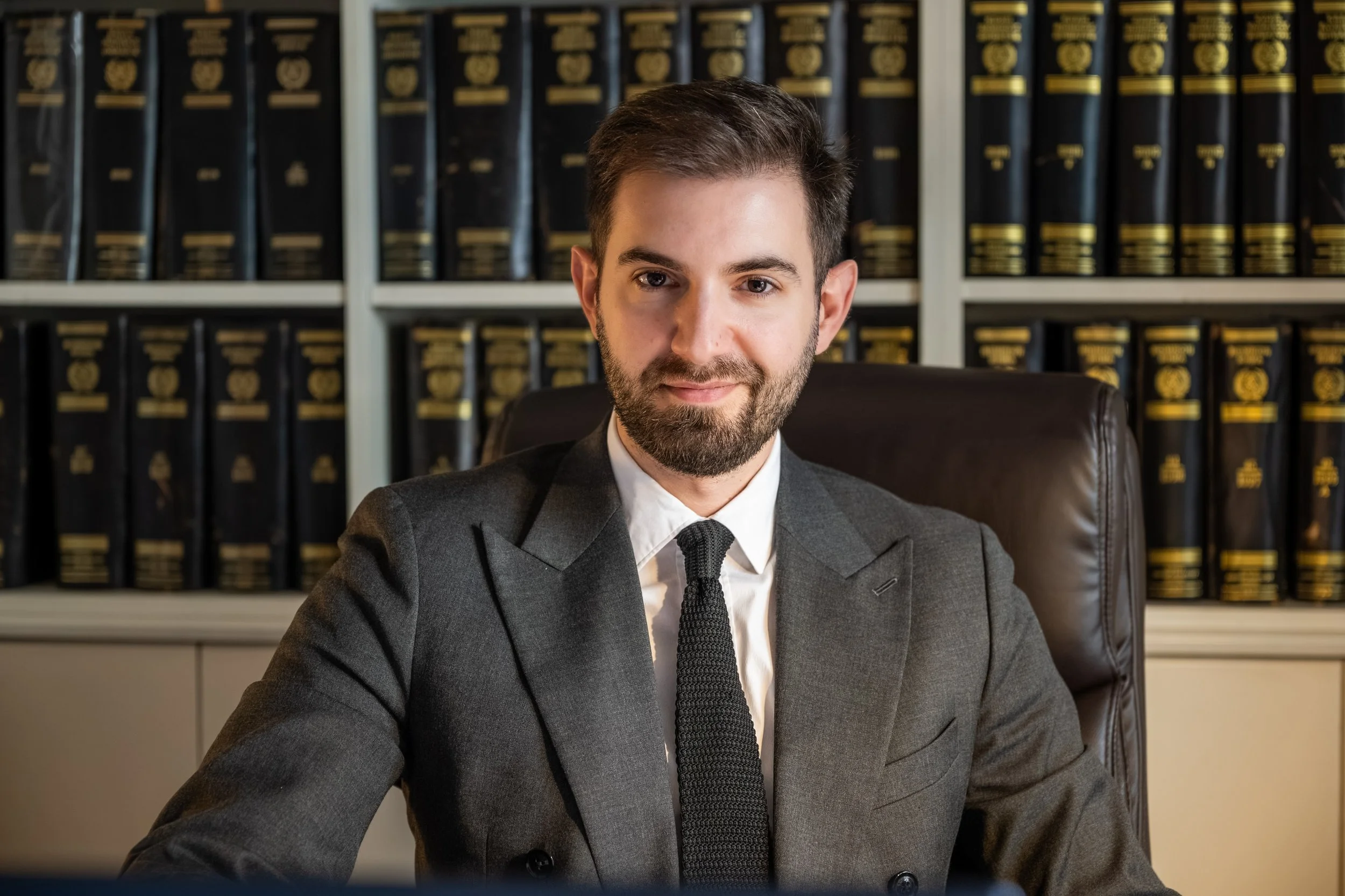 A man with brown hair, beard, wearing a dark suit and tie, sitting at a desk in front of a bookshelf filled with legal books.