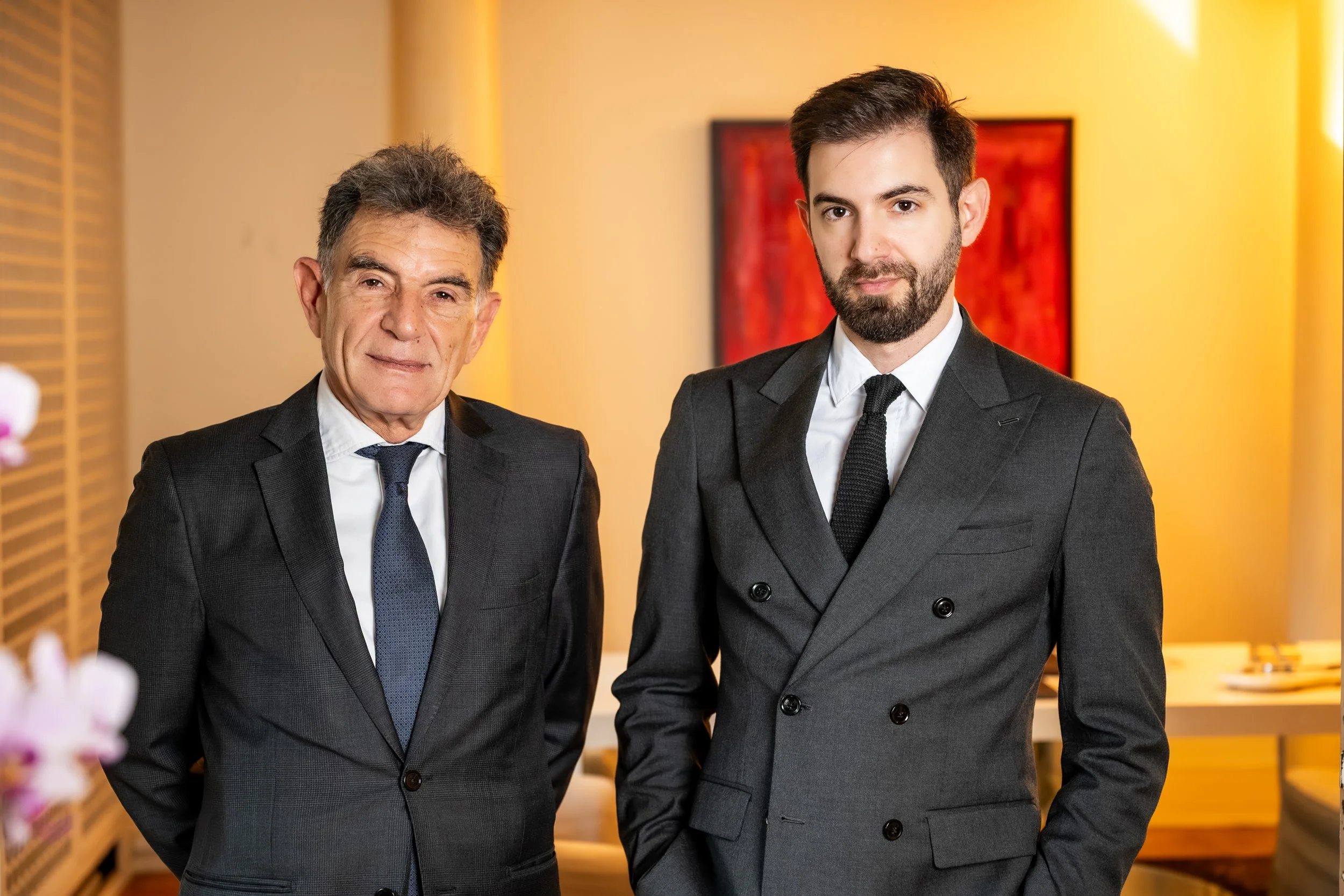 Two men dressed in suits standing in an indoor setting, one older with gray hair and the other younger with dark hair and a beard, posing for the photo.