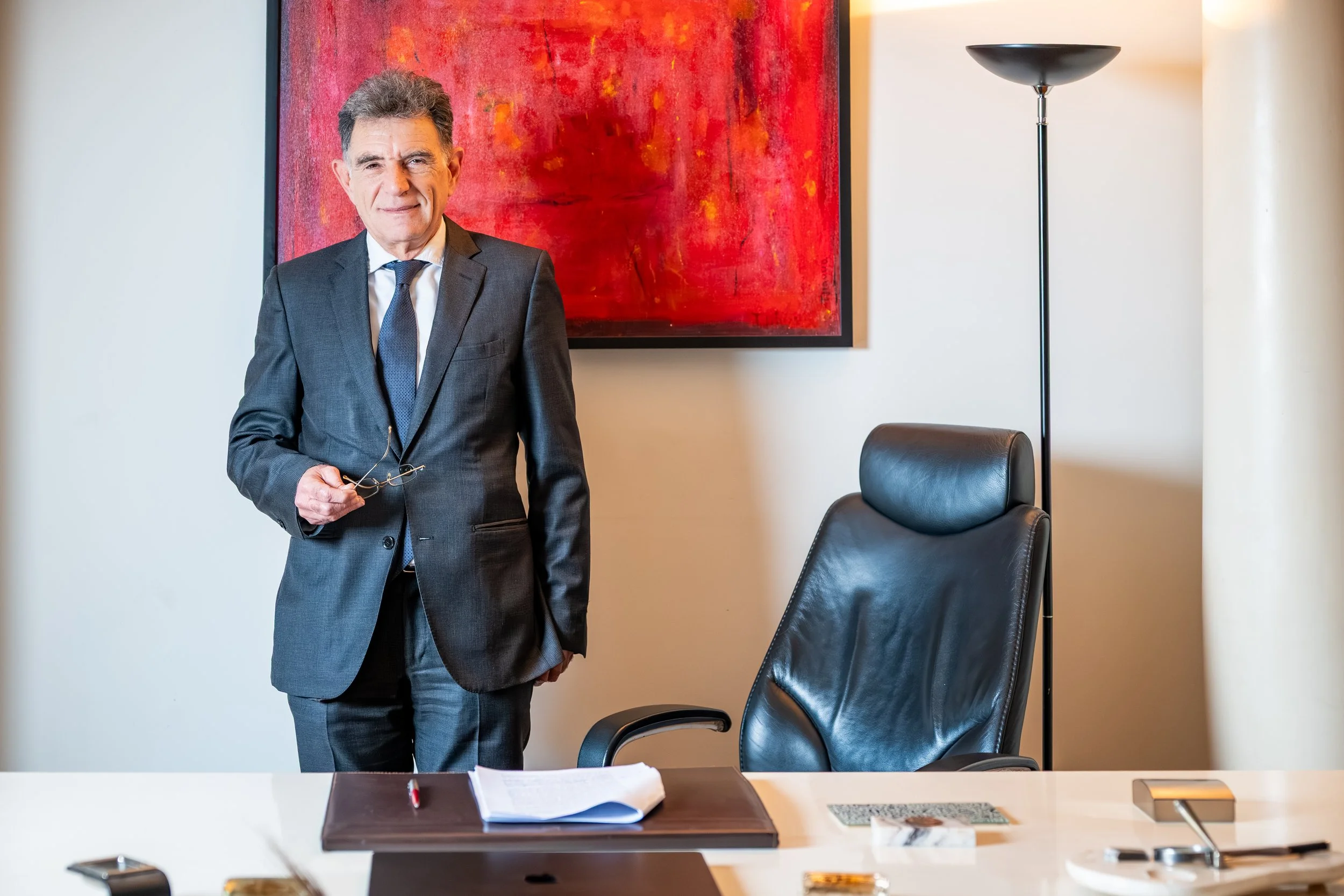 A man in a gray suit holding glasses standing in a modern office with a red abstract painting on the wall, an office chair, a white table with papers, a pen, and a smartphone.