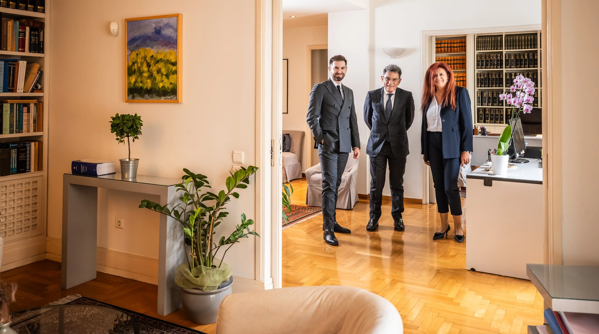 Three professionally dressed people standing in an office, smiling for the camera, with bookshelves and a desk with a computer in the background.
