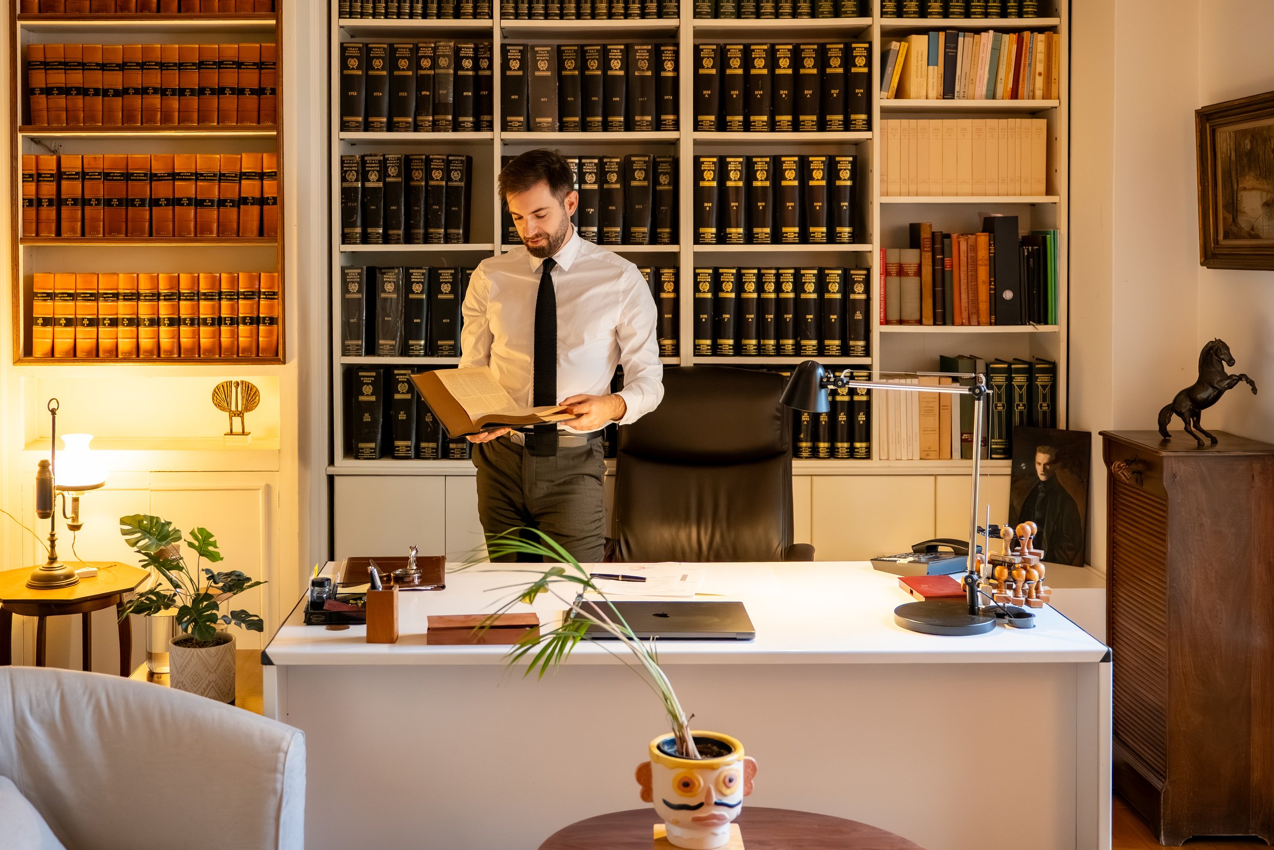 A man in a white shirt and black tie standing in an office filled with bookshelves, reading a book. The office has a desk with a lamp, plants, and decorative items including a chess set and a horse sculpture. The background includes bookshelves filled with legal books and other books.