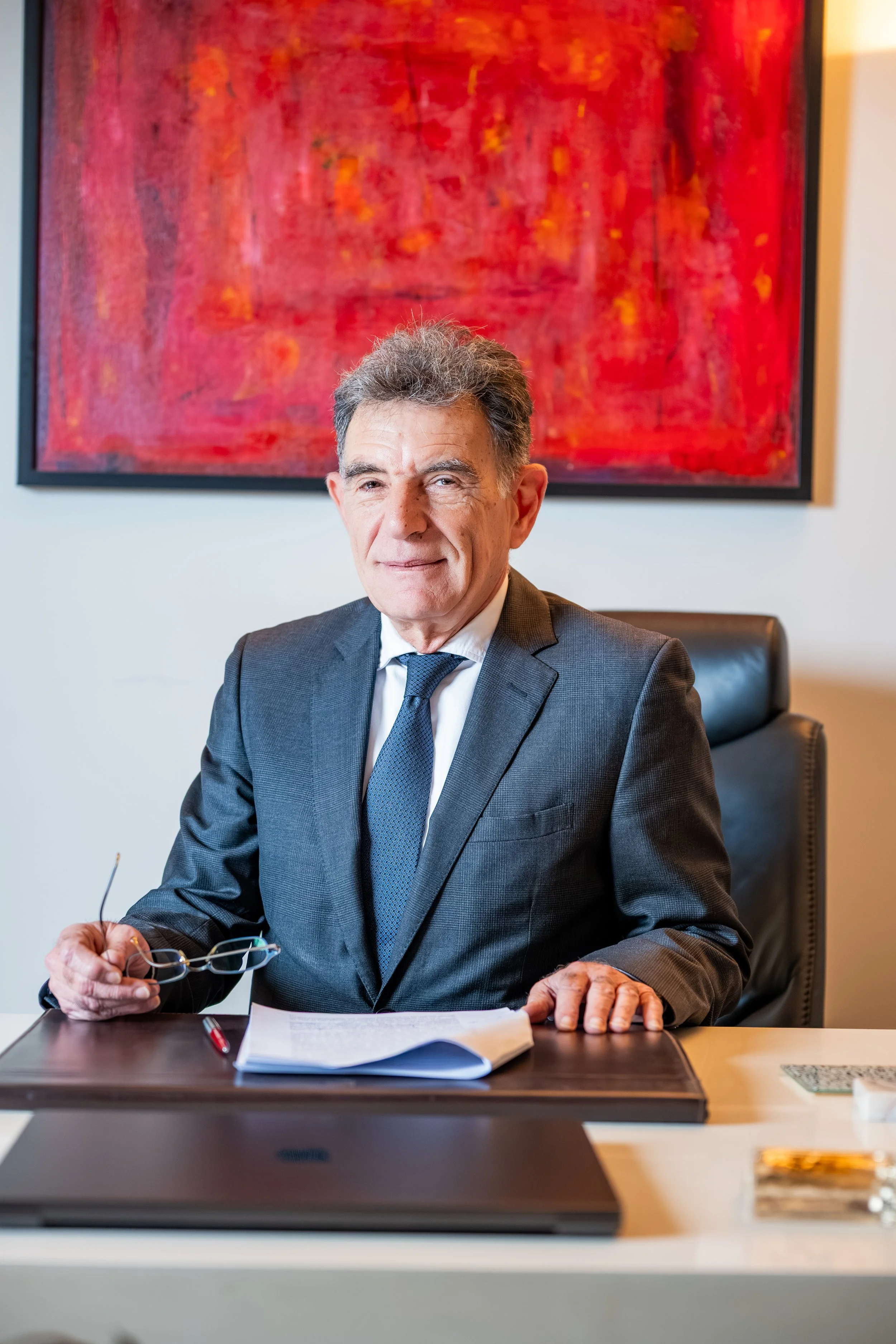 A senior man with gray hair and a dark suit sitting at a desk in an office, holding glasses in his right hand. There are papers, a pen, and a mask on the desk. A large red and orange abstract painting hangs on the wall behind him.