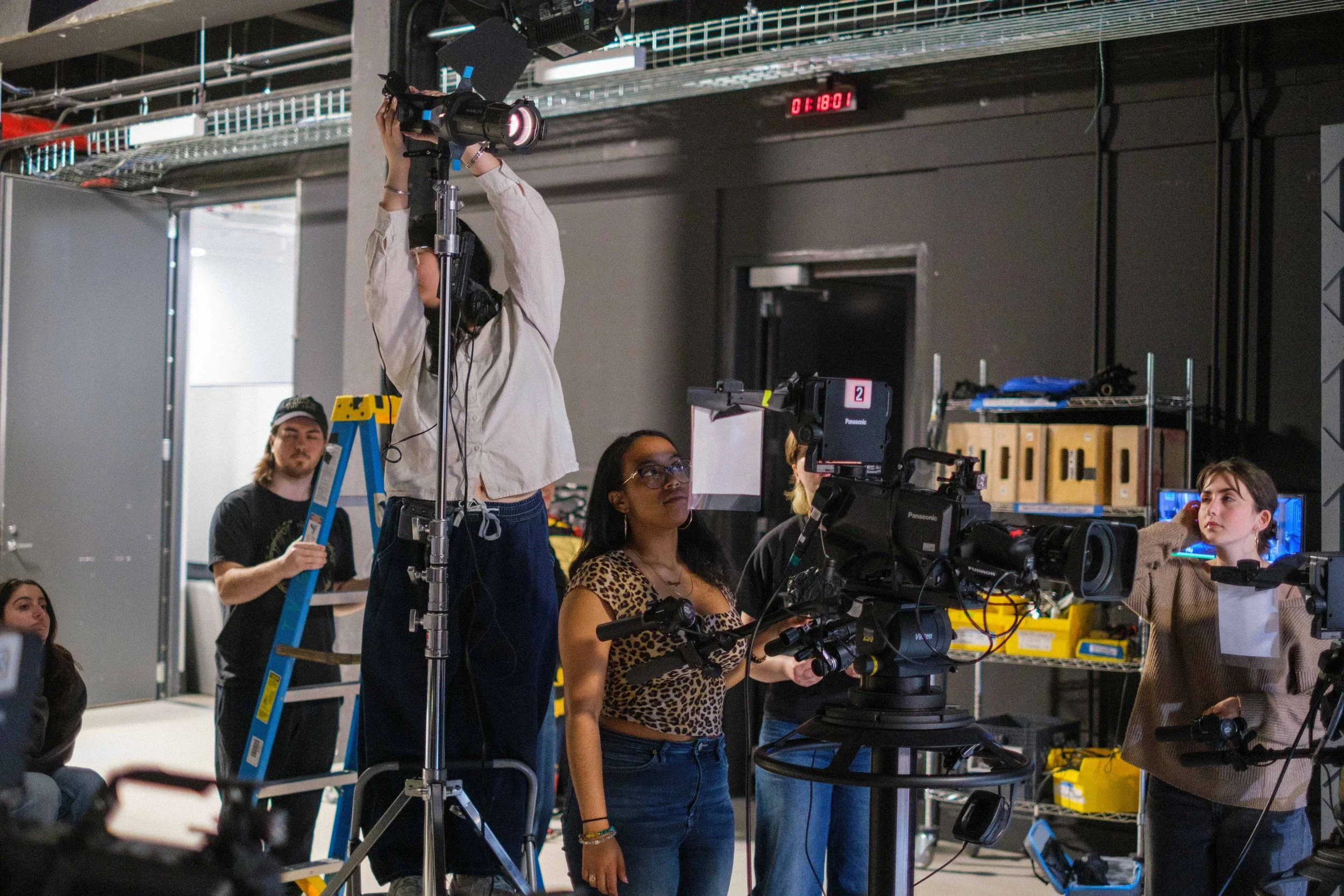 People working in a film or video production studio with cameras, lights, and equipment, while a woman looks through a camera viewfinder.