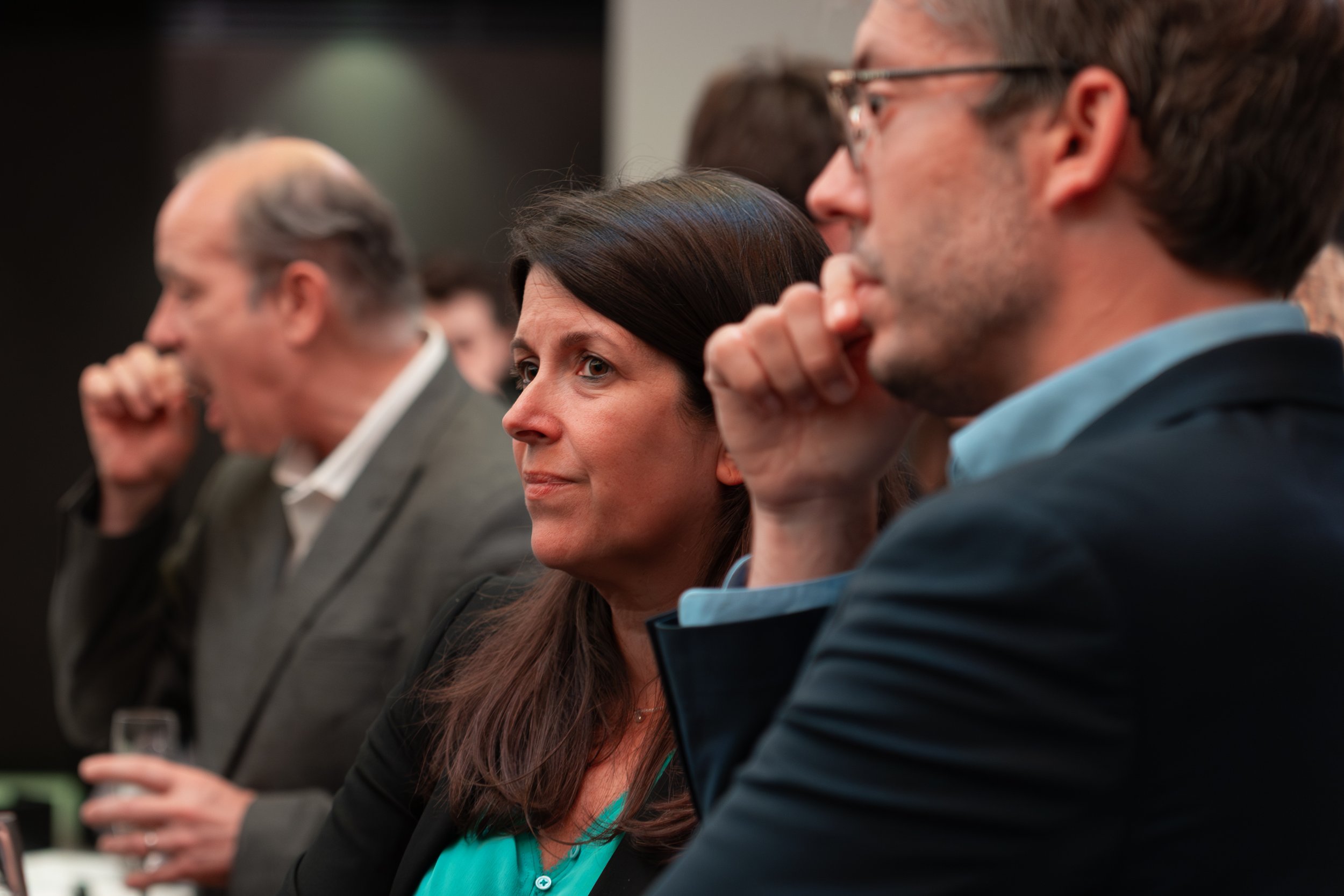 Groupe de personnes assises en rangée lors d'une conférence ou d'une réunion, écoutant attentivement, avec une femme au centre regardant devant et un homme avec lunettes à droite en train d'écouter.