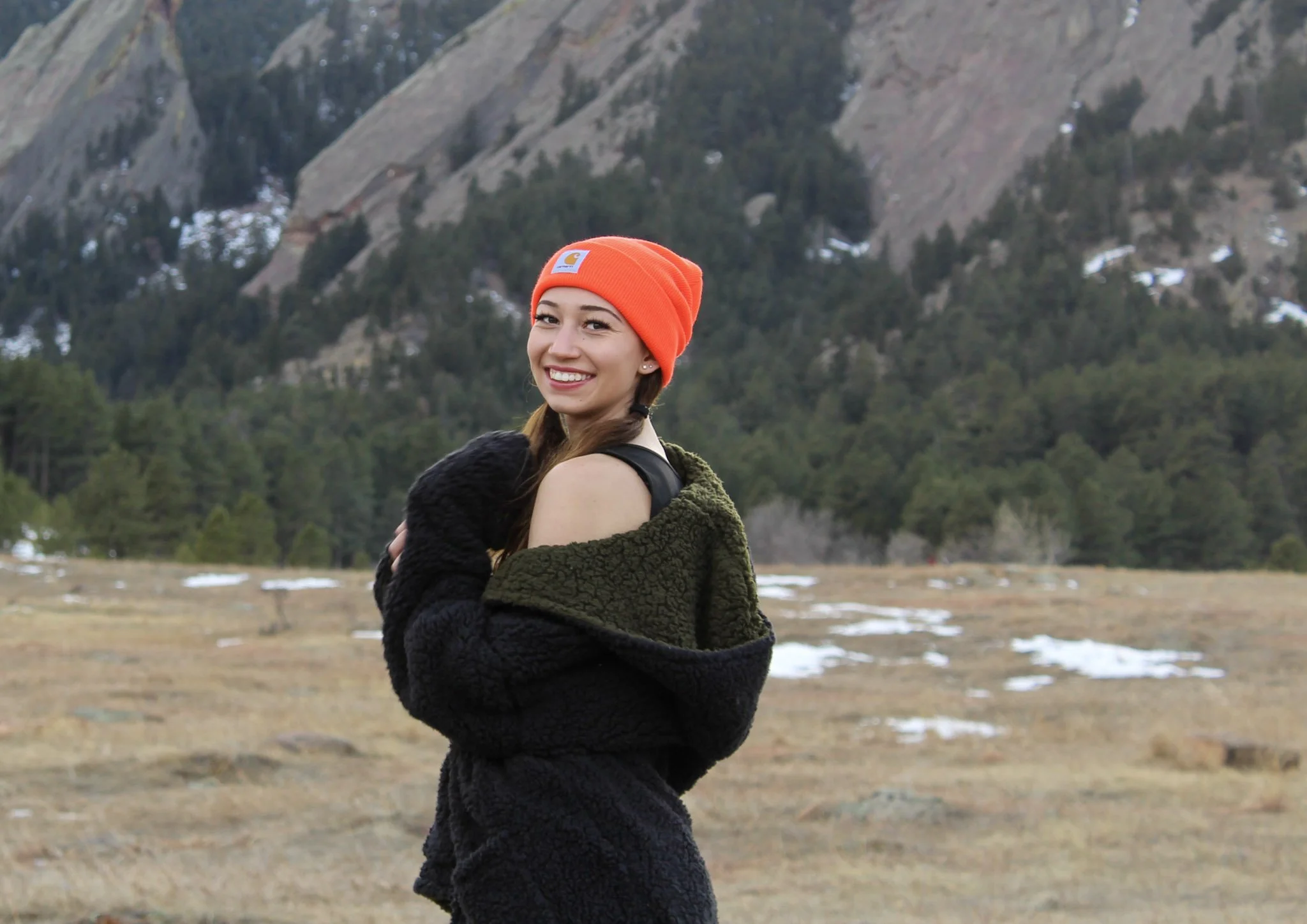 A young woman smiling outdoors, wearing an orange beanie and a black and green jacket, with mountains and trees in the background.