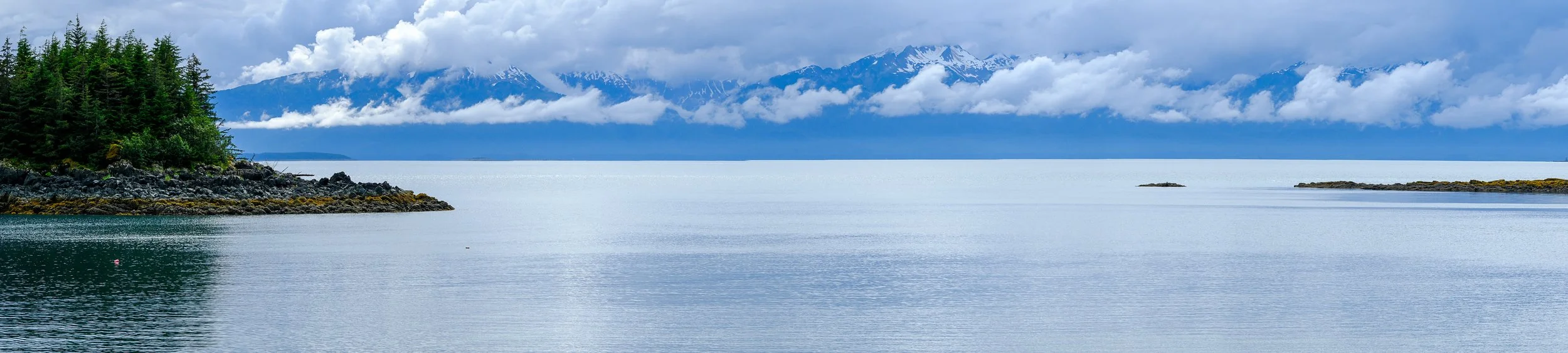 A calm body of water surrounded by green pine trees and rocky shores, with snow-capped mountains and clouds in the background.
