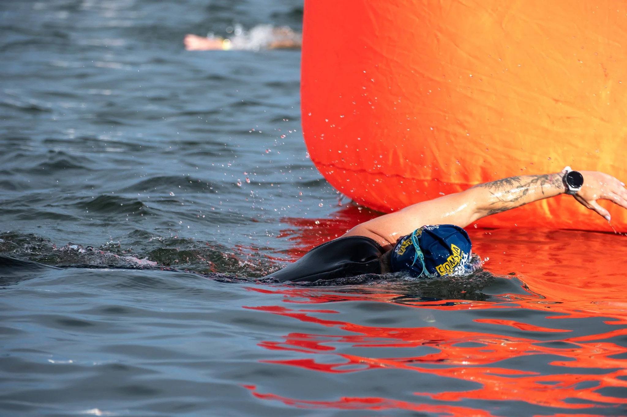 A swimmer in a black wetsuit and blue swim cap is performing a freestyle stroke in choppy water next to an orange safety buoy.