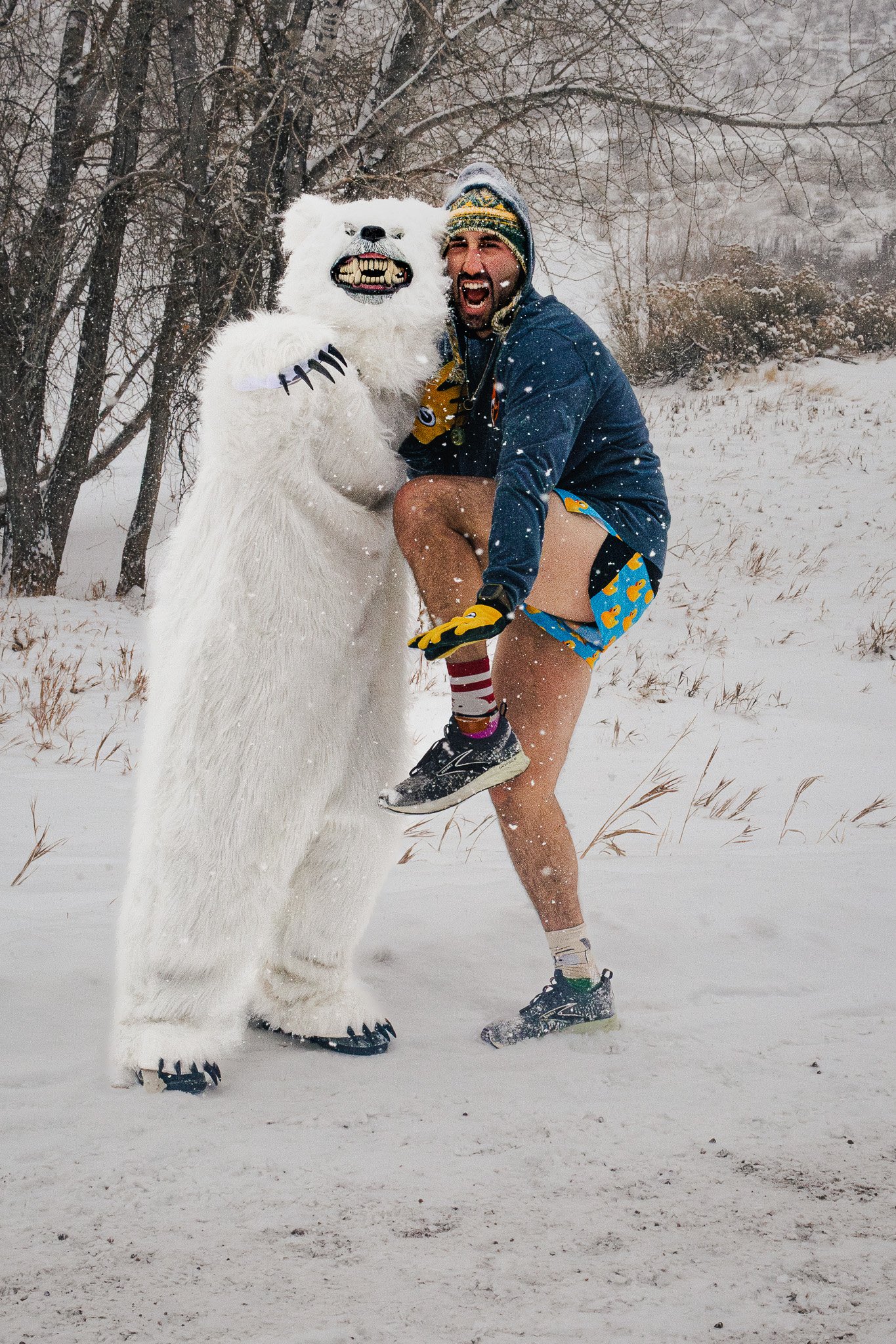 A man wearing a colorful beanie, shorts with rubber ducks, a blue jacket, and gloves, is playfully posing next to a large, anthropomorphic polar bear costume with a snarling face, in a snowy outdoor setting with trees and snow-covered ground.