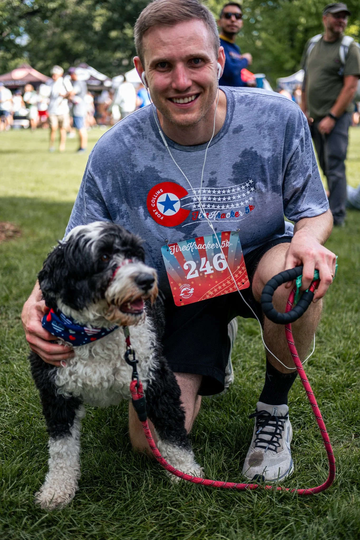 A man smiling and crouching on grass at a park or outdoor event with a black and white dog, both wearing red, white, and blue patriotic bandanas. The man has short, light brown hair, wears a gray T-shirt with a race bib, and earphones. The background
