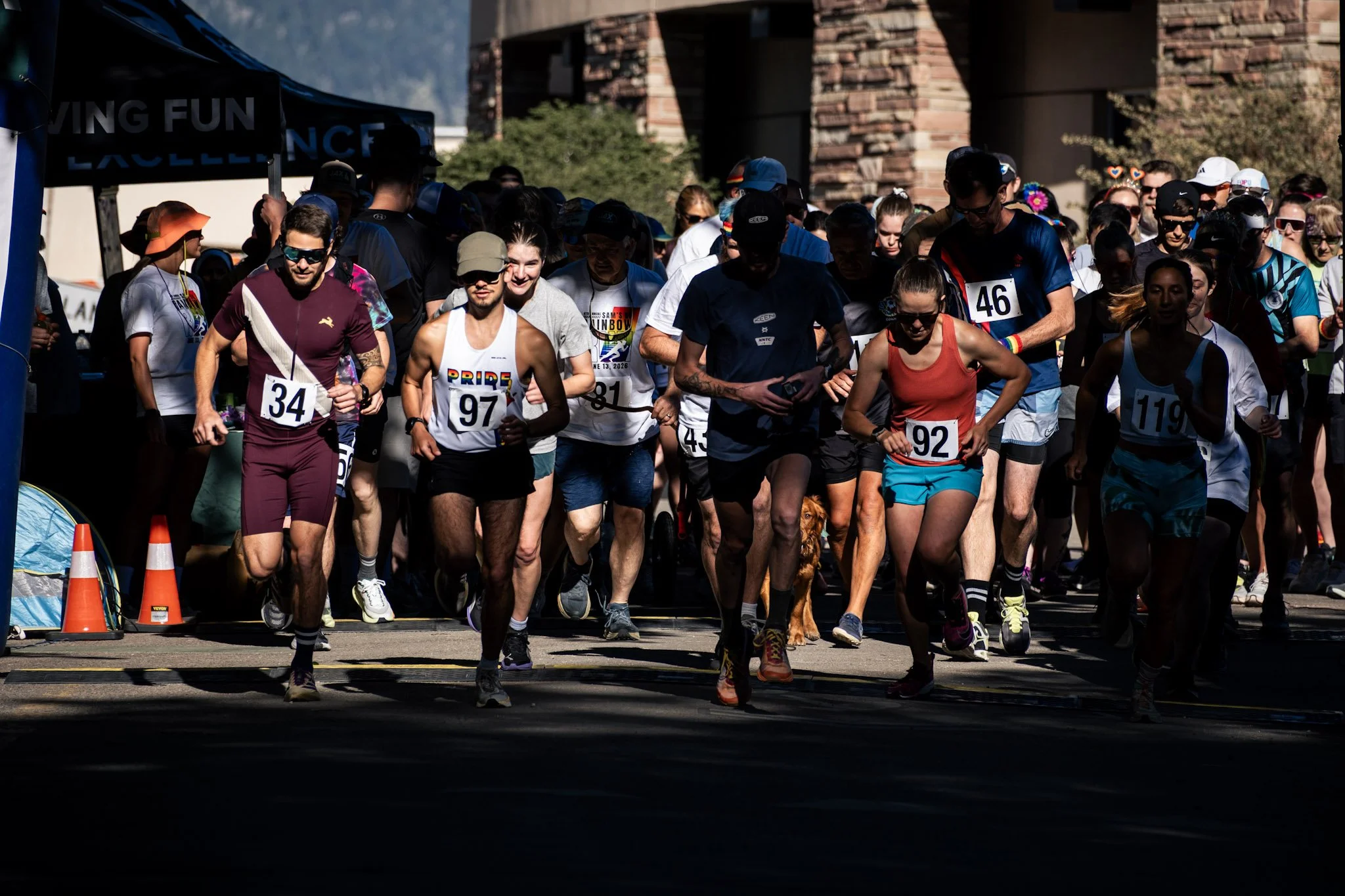 Group of runners at the start of a race, wearing race bibs and athletic clothing, with some smiling and others focused, under a tent with orange traffic cones in the foreground.