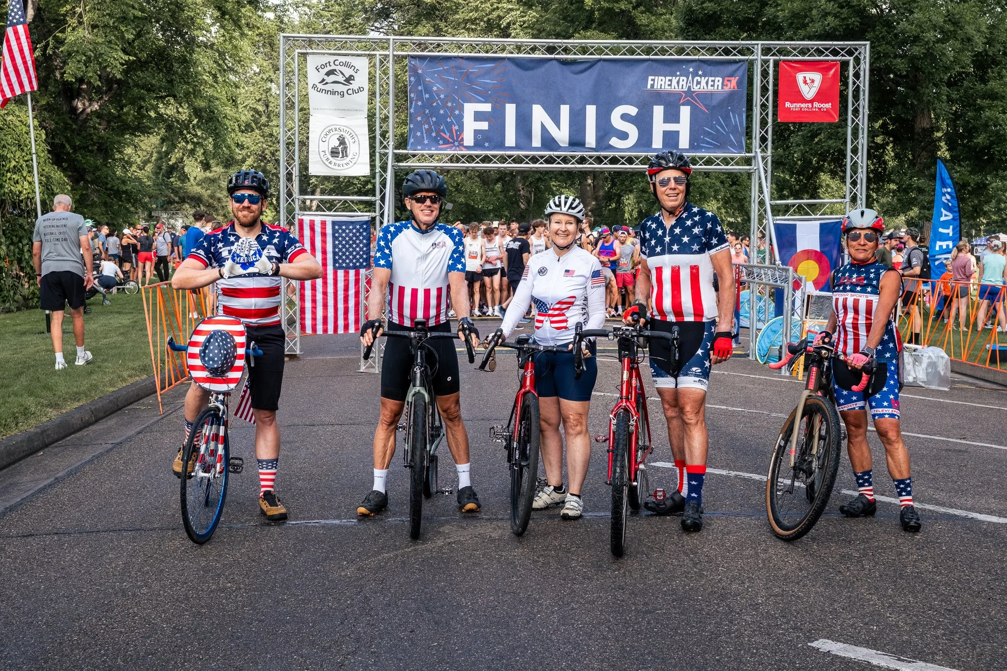 Six cyclists dressed in red, white, and blue patriotic outfits standing with bikes at the finish line of a race, with a crowd of people and trees in the background.