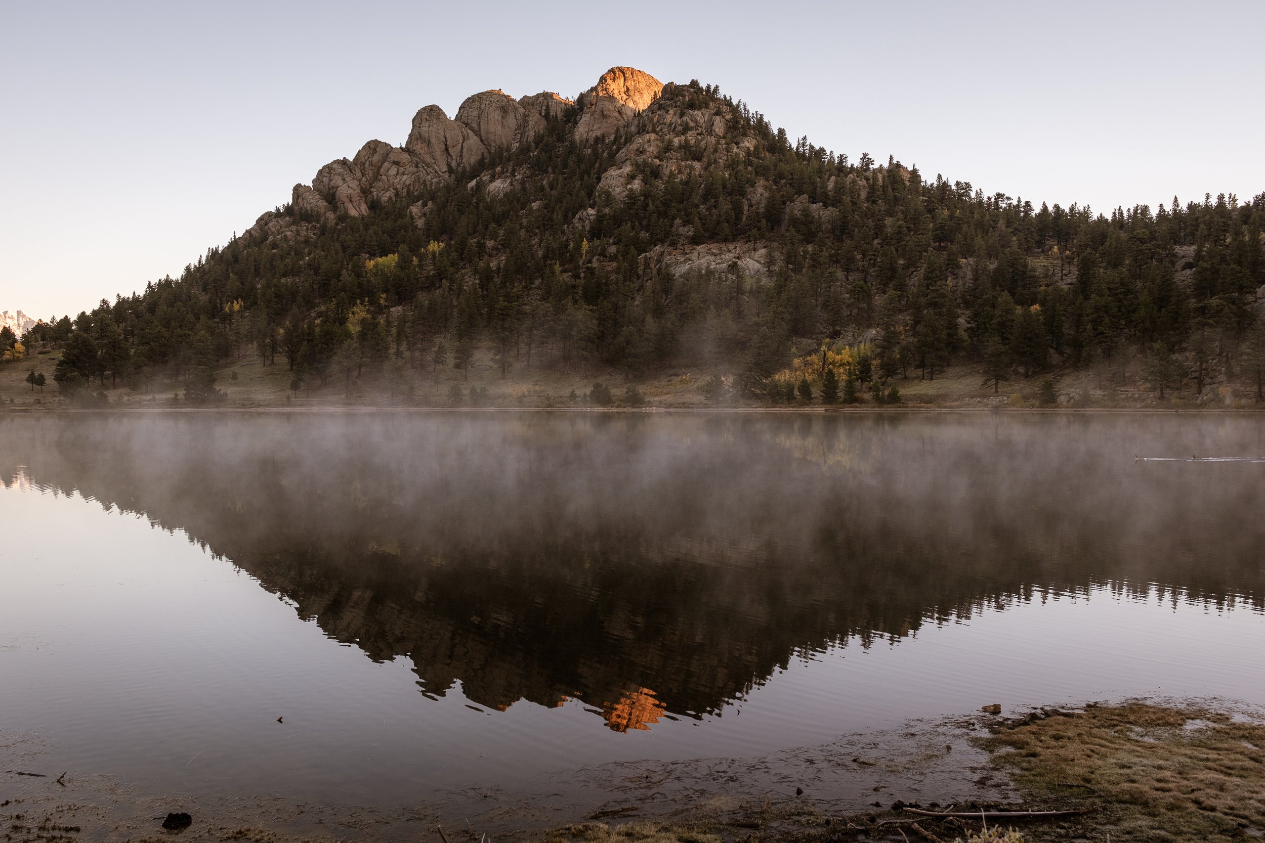 A mountain with rocky peaks reflected in a calm body of water, surrounded by a forested shoreline with mist rising from the water.
