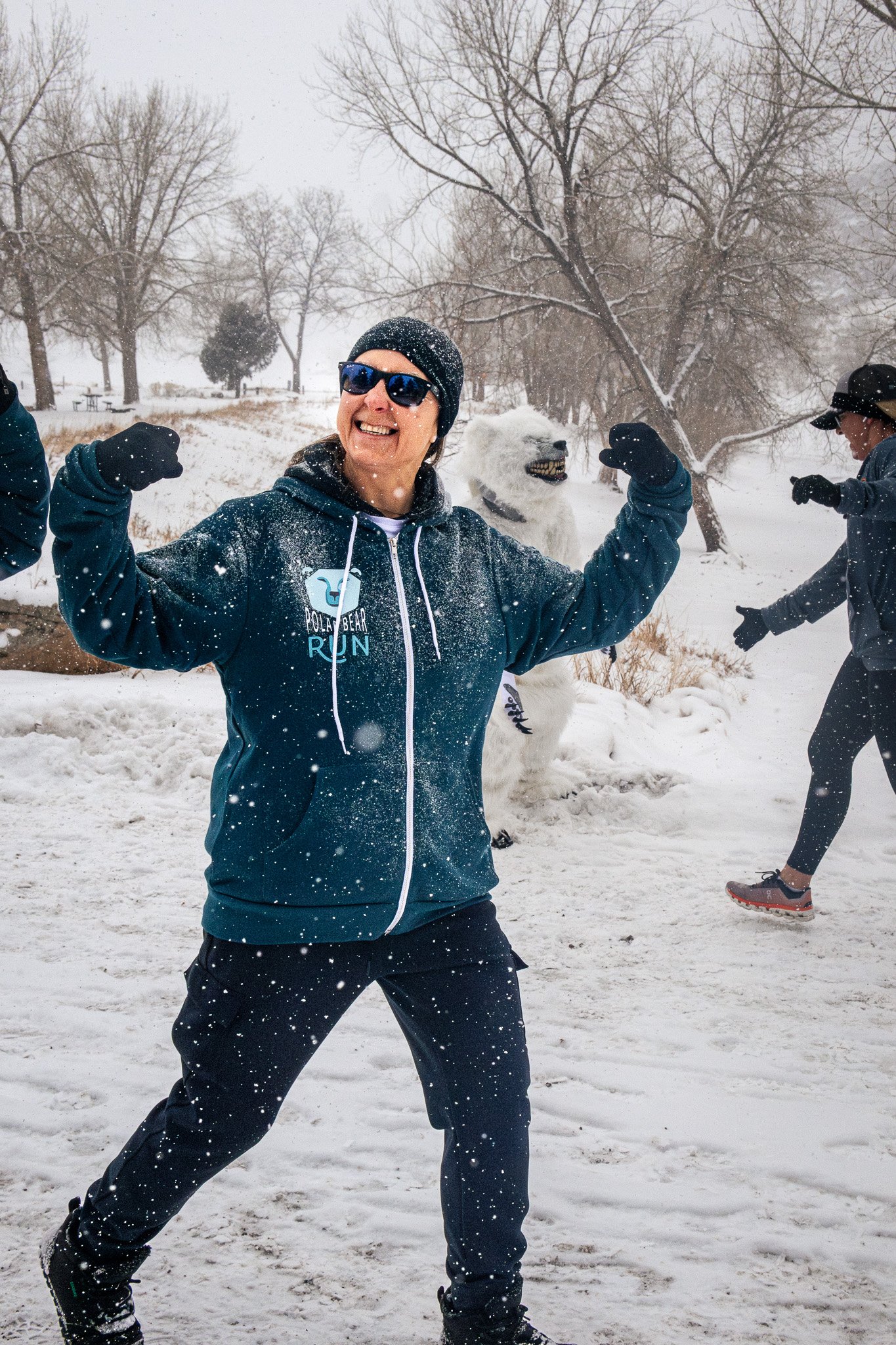 A person wearing sunglasses, a beanie, and a blue jacket poses with flexed arms in a snowy park, with a white dog in the background.