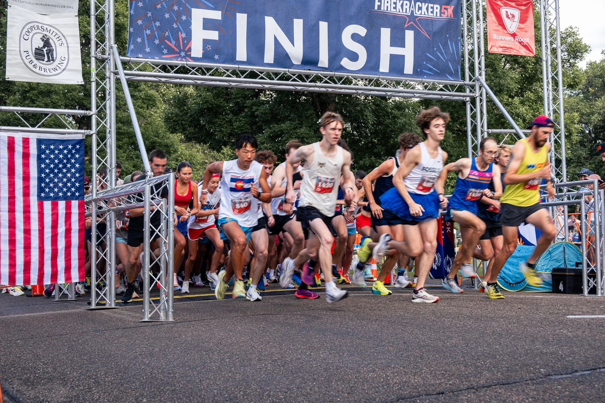 A group of runners start a race at the starting line under a large decorated finish banner with flags and sponsors, outdoors amidst trees.
