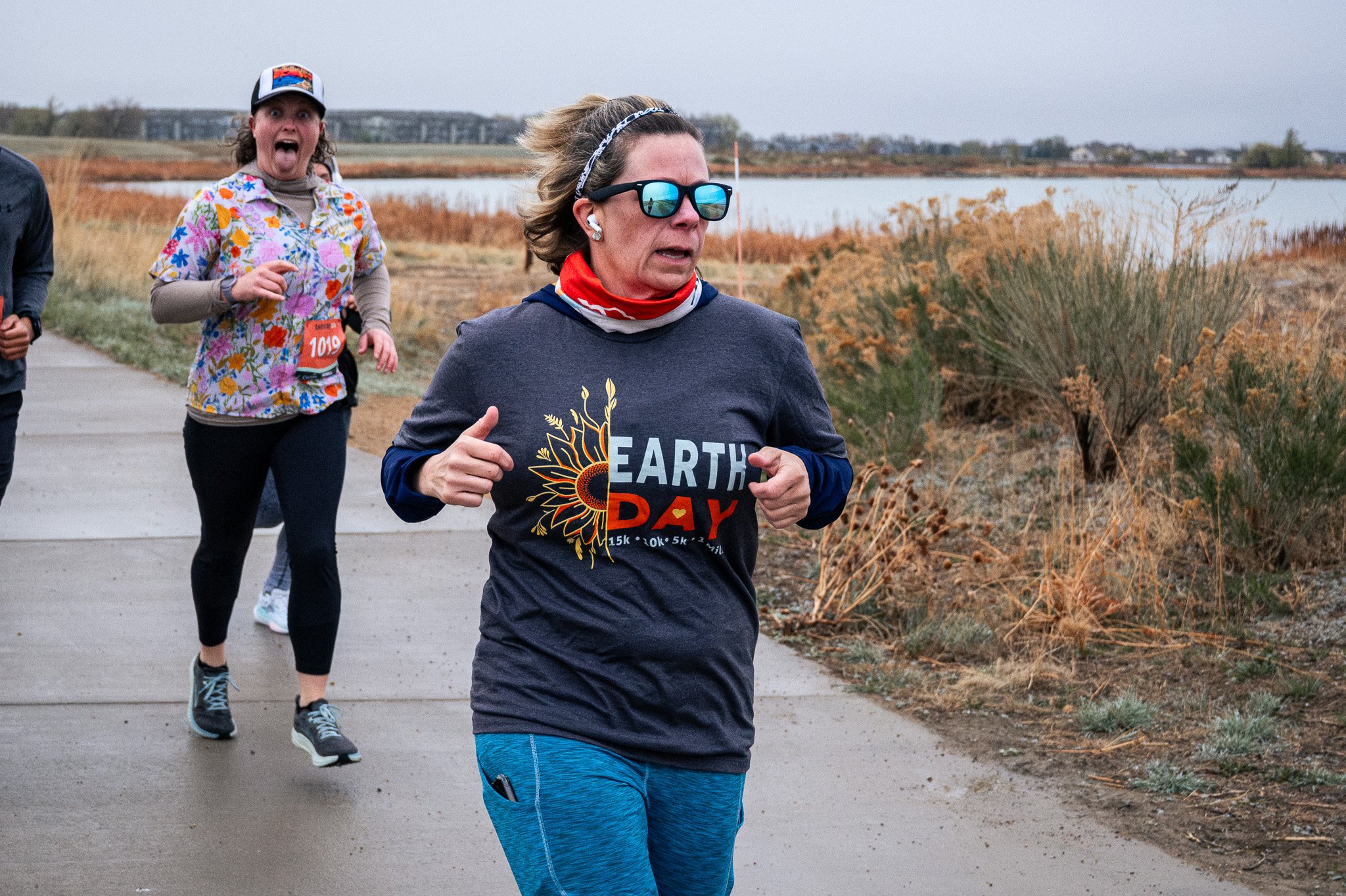 A group of people running outdoors on a cloudy day near a water body with brown grass and bushes.