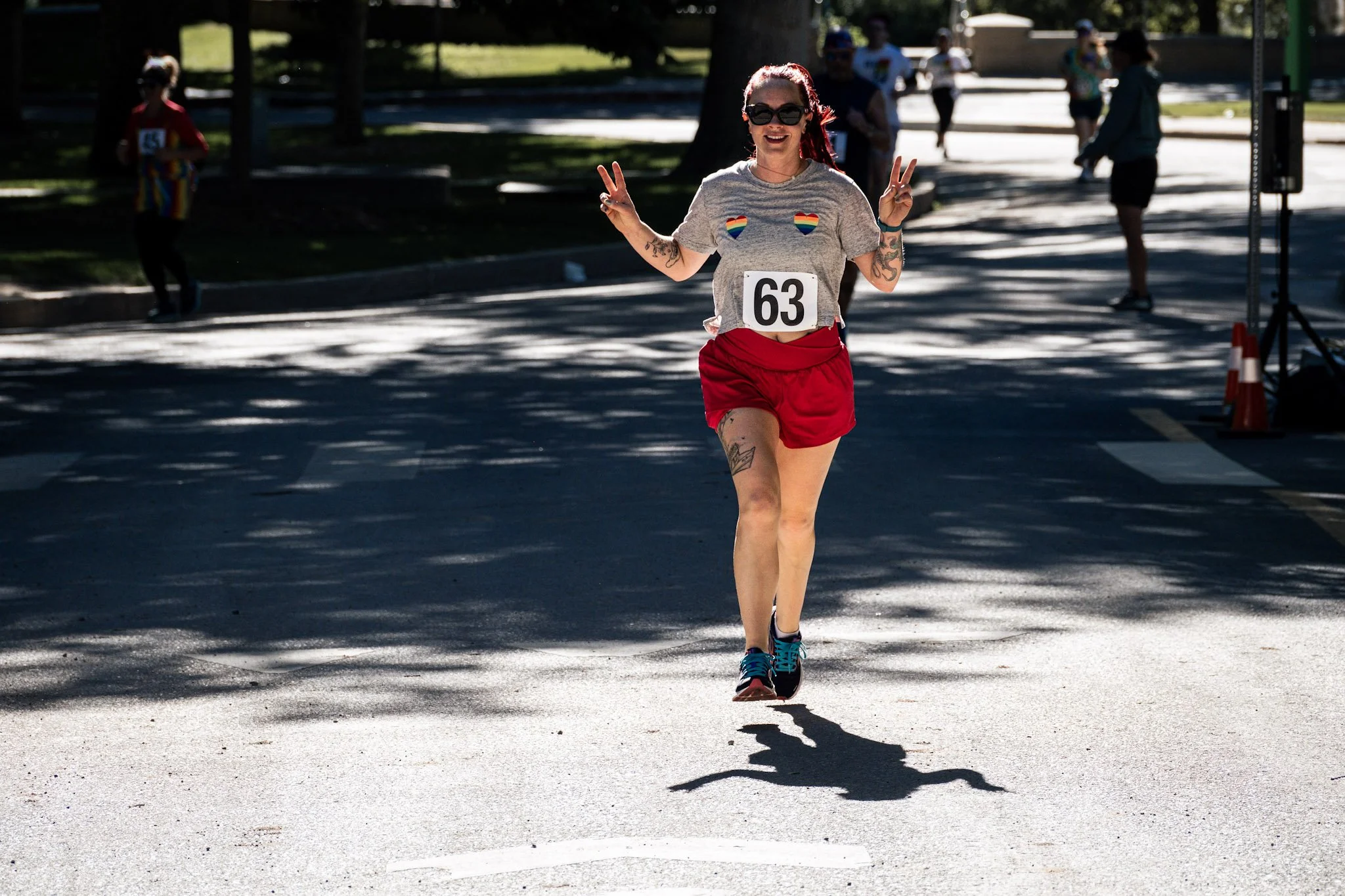 A woman running in a race, wearing a gray T-shirt with rainbow heart patches and a number 63, red shorts, sunglasses, and smiling while making peace signs with both hands, with other runners and trees in the background.
