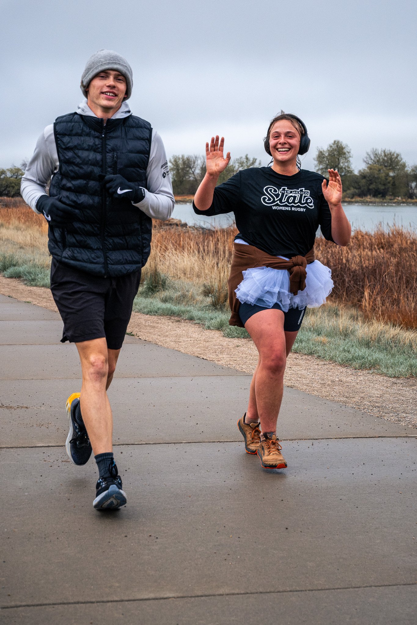 Two people jogging outdoors along a paved path near a body of water, with trees in the background. One is a young man wearing a grey beanie, black vest, and black shorts. The other is a young woman with headphones, wearing a black T-shirt that says '