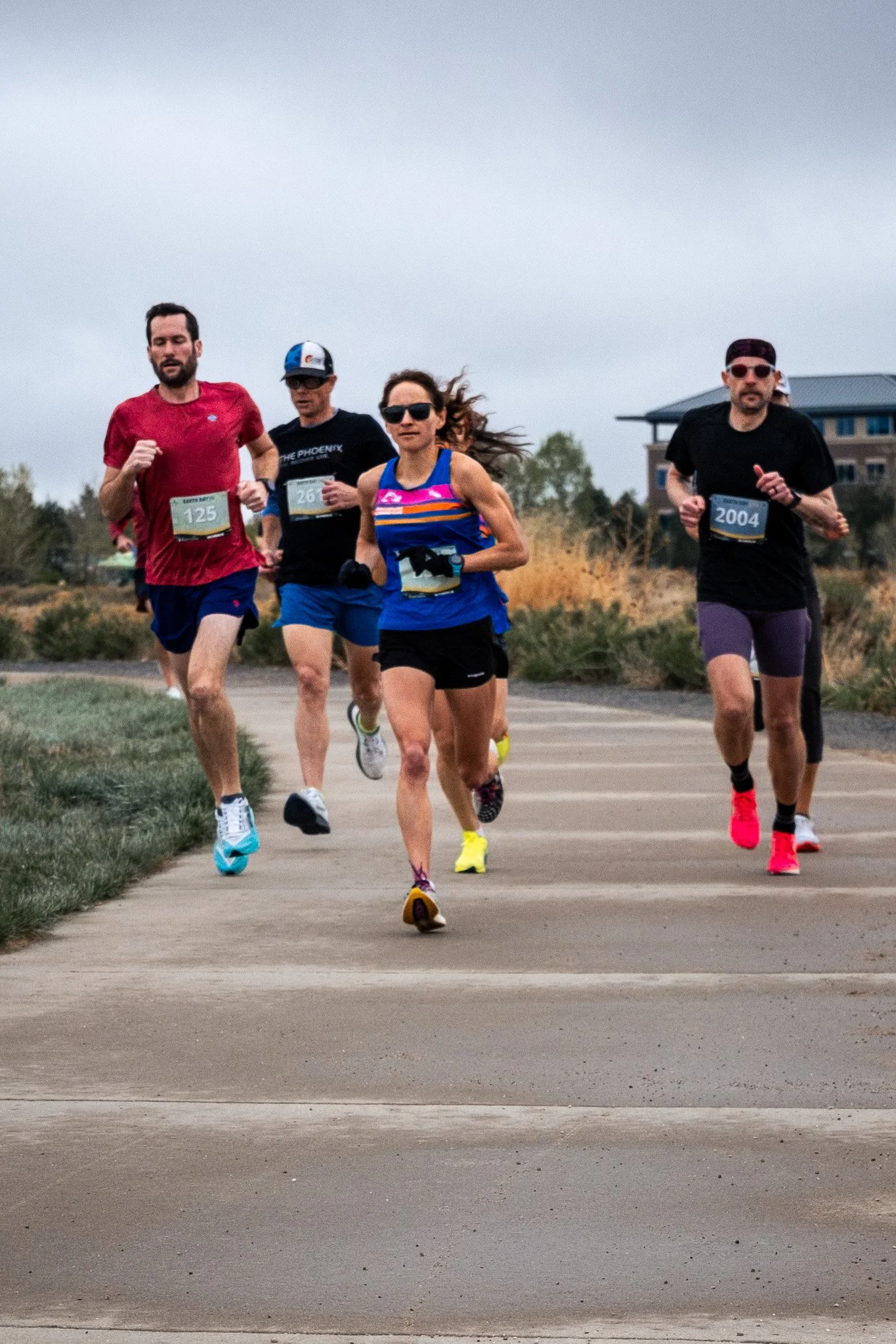 A group of five runners participating in a race outdoors, dressed in athletic clothing and running on a paved path, with a house and trees in the background on a cloudy day.