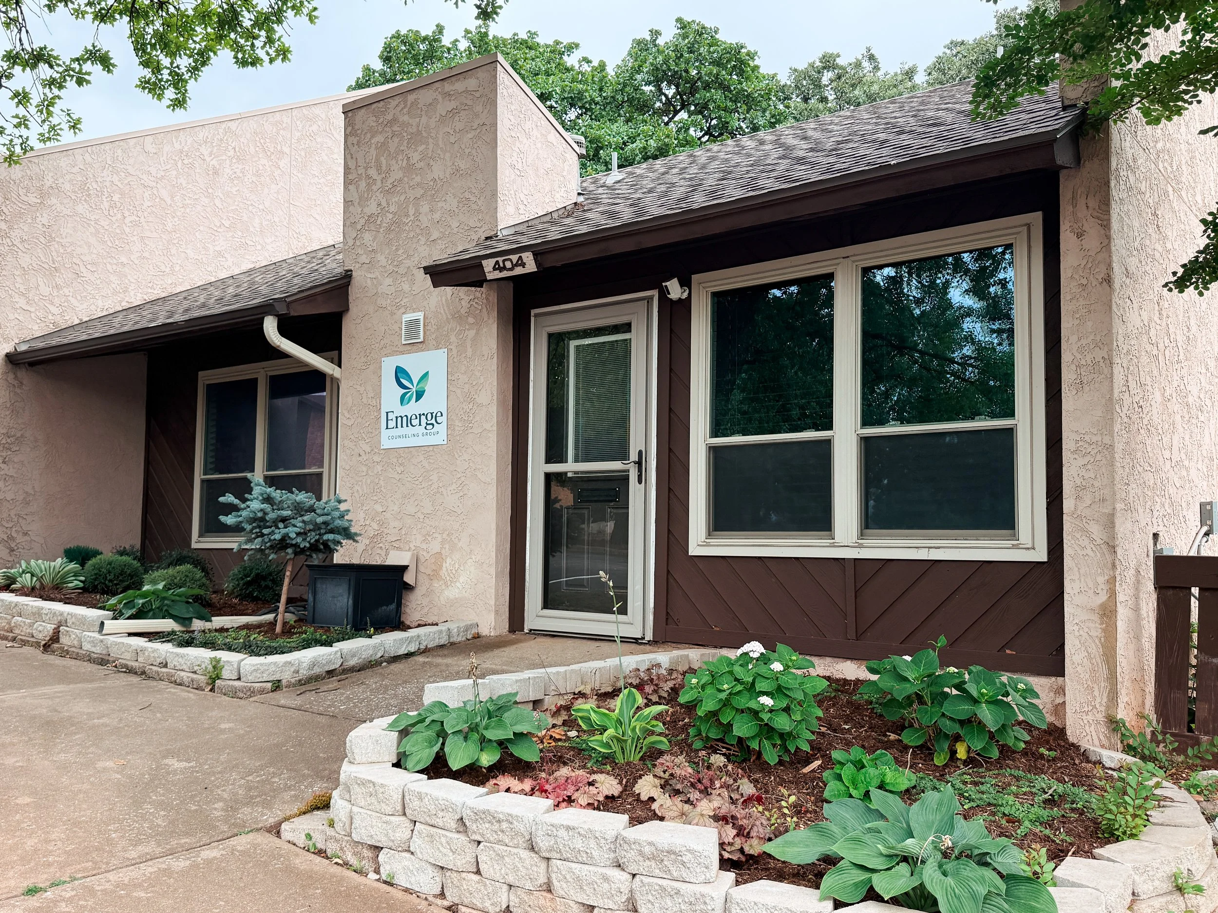 Front view of a beige stucco residential building with a dark brown wooden trim, a large window, and a white door. The building number '404' is above the door, and there is a sign that reads 'Emerge Counseling Group' with a leaf logo. In front of the house, there is a landscaped garden with various green plants and a small Christmas tree, bordered by a white stone edging, and a concrete sidewalk.
