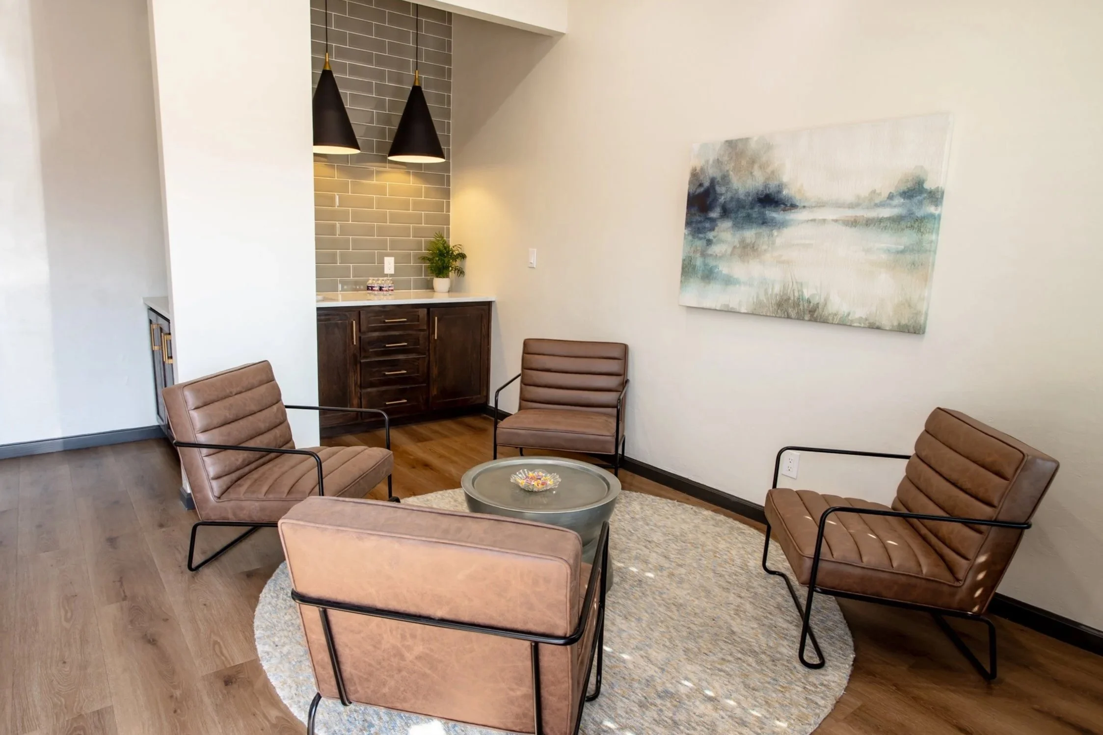 A waiting area with four brown leather chairs with black metal frames arranged around a round coffee table on a beige rug. A small sideboard with a potted plant and supplies is against the wall, with two black cone-shaped pendant lights hanging overhead. A large abstract landscape painting decorates the white wall.