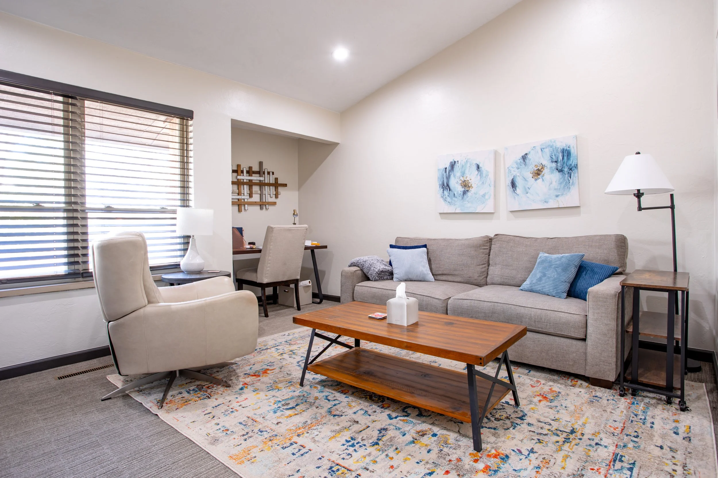 Living room with a beige armchair, a couch with blue and gray pillows, a wooden coffee table, a colorful area rug, wall art, a lamp, and windows with dark blinds.