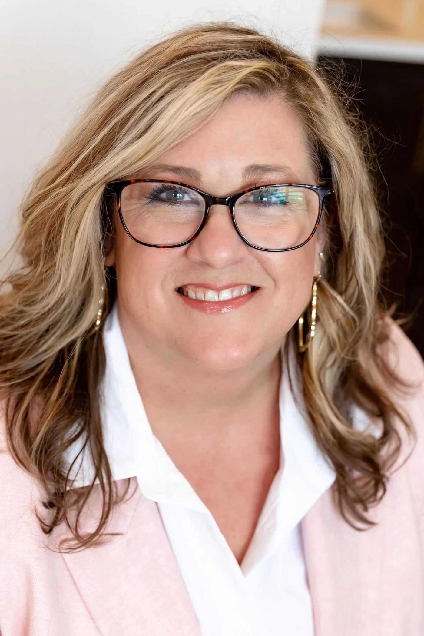Close-up of a middle-aged woman with blonde hair, wearing glasses, smiling, dressed in a white shirt and pink blazer.