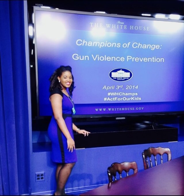 Gianna Judkins standing in front of a large screen at the White House, which displays a presentation titled 'Champions of Change: Gun Violence Prevention' dated April 3rd, 2014, with hashtags #WHChamps and #ActForOurKids.