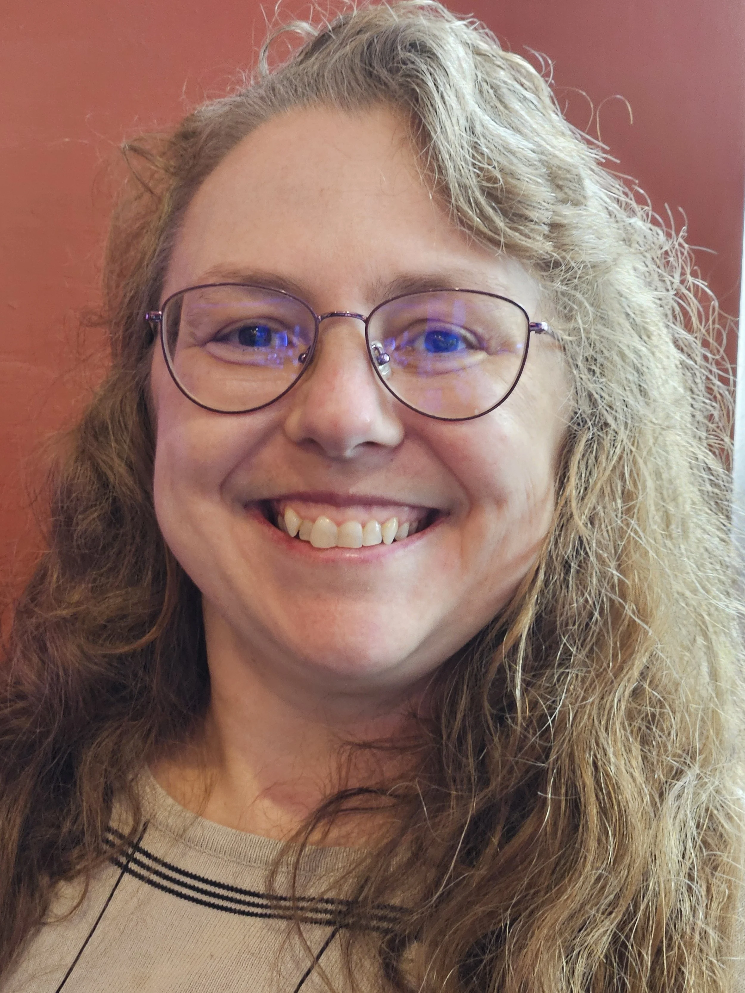 Close-up of a smiling woman with curly light brown hair, wearing glasses, against a reddish background.
